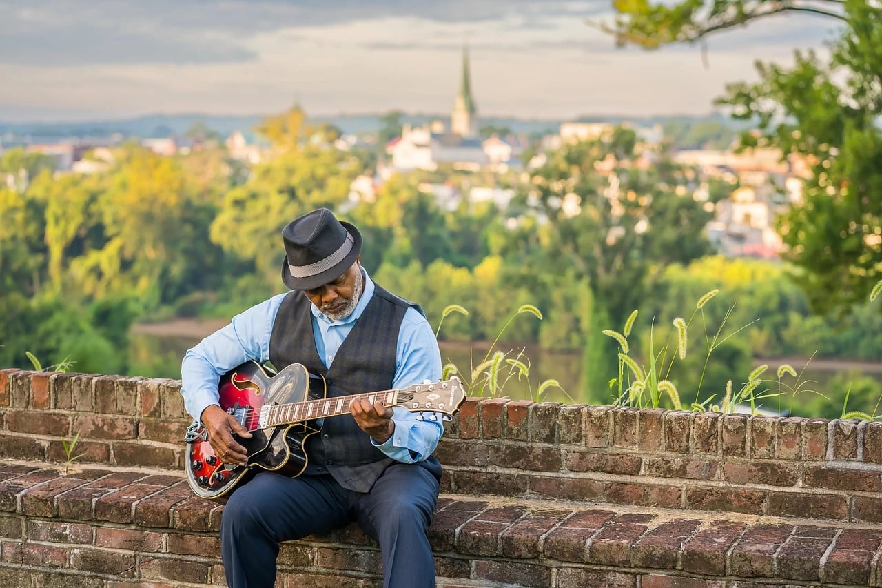 Older man in a fedora and vest playing a red electric guitar on a brick wall overlooking a town