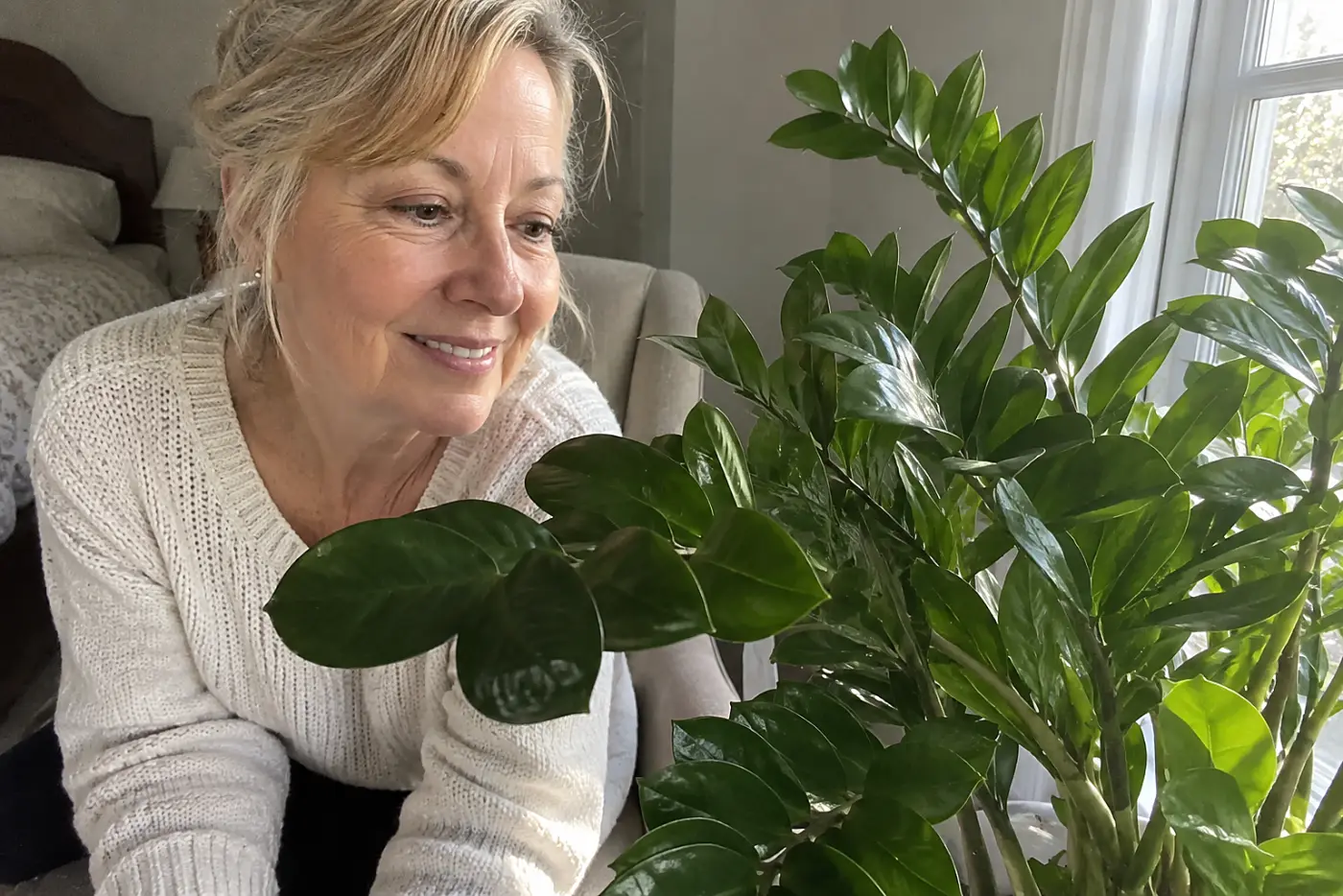 A smiling woman in her sixties wearing a white sweater admires a green ZZ plant.