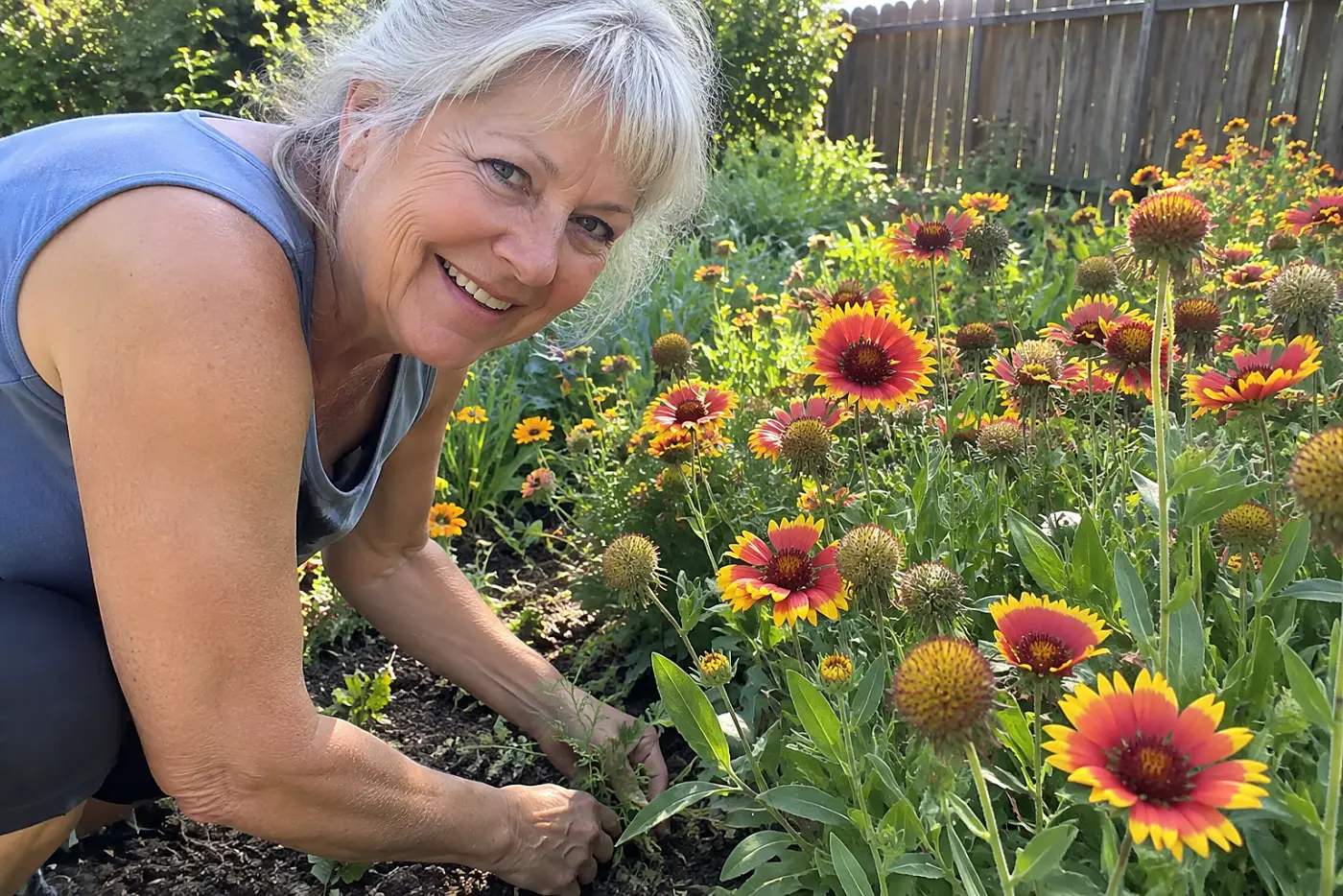 A woman in her sixties smiles while tending blanket flowers in a sunny garden bed.