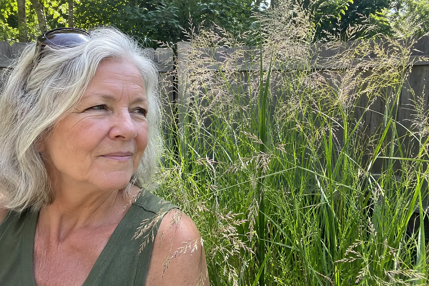 A woman in her sixties wearing a green top stands near tall switchgrass along a fence.
