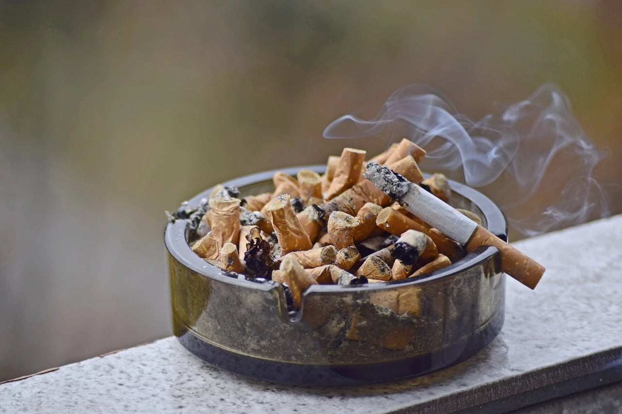 Glass ashtray overflowing with cigarette butts and a lit cigarette with smoke rising, on a stone ledge