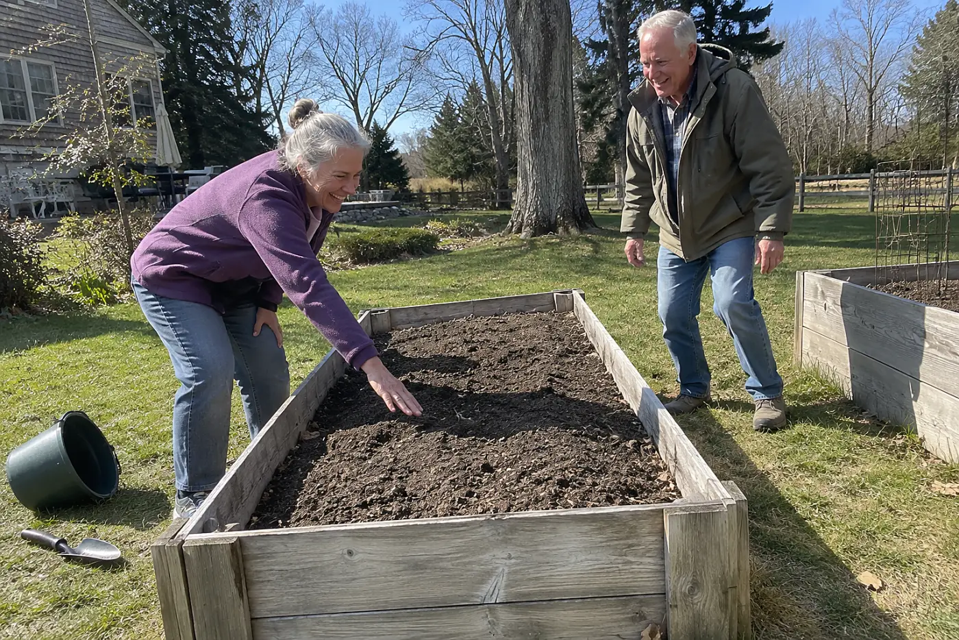 Older woman reaching toward the middle of an empty raised bed in her yard.