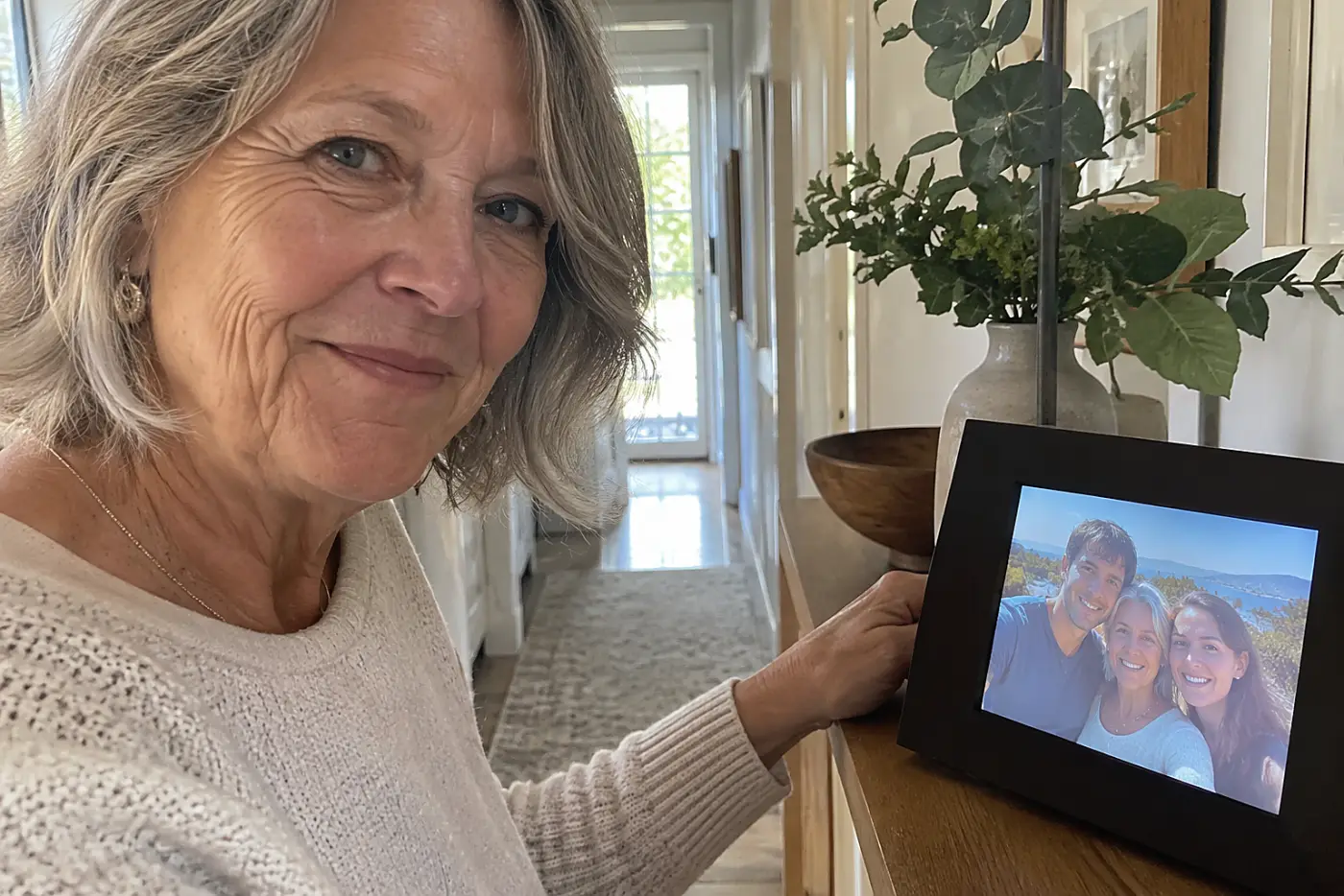 An older woman watches a new family photo appear on a digital frame on her hallway shelf.