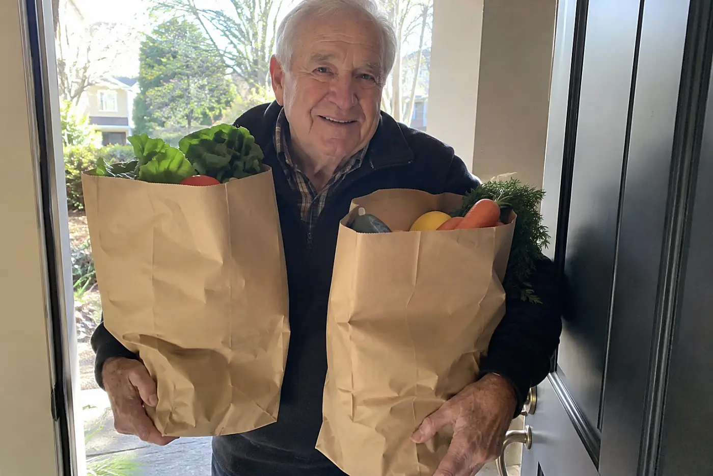 A grandfather carries delivered grocery bags from his front porch into the house.