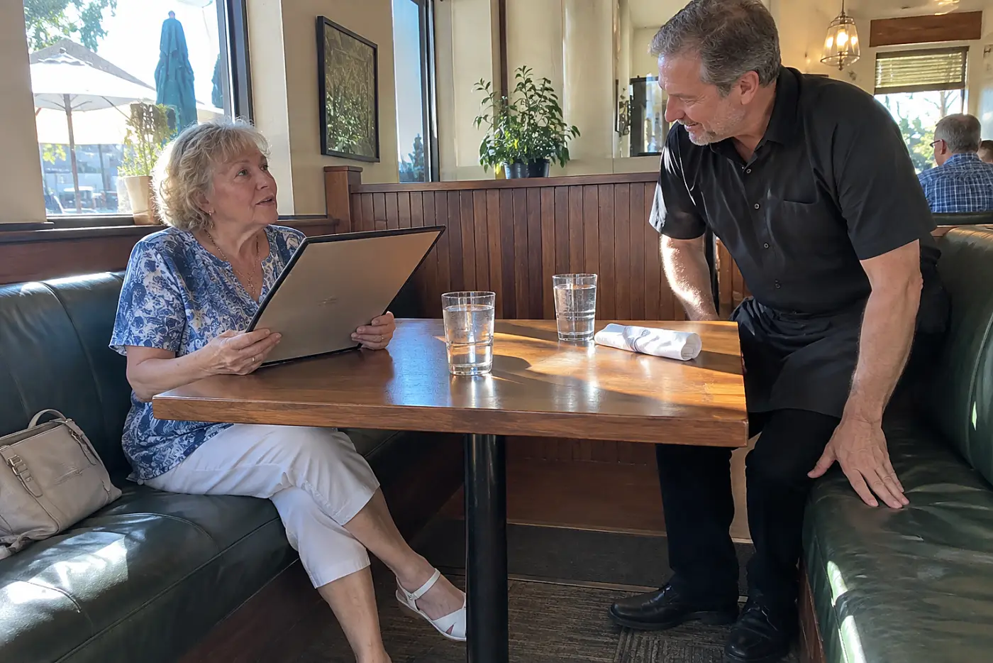 Older woman reading a menu across from a server in a restaurant booth.