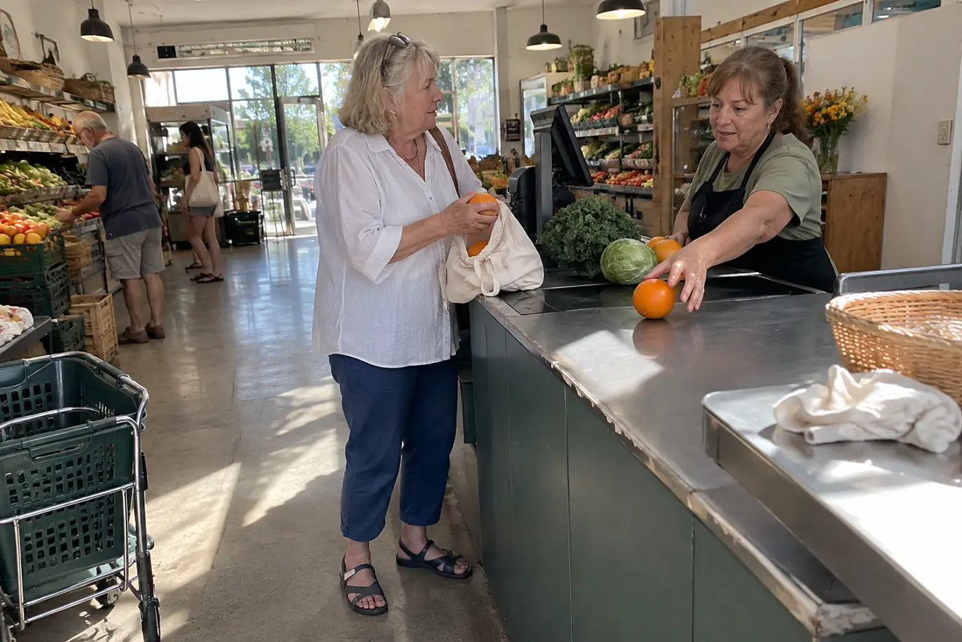 Older woman speaking to a cashier while bagging oranges at a grocery store.
