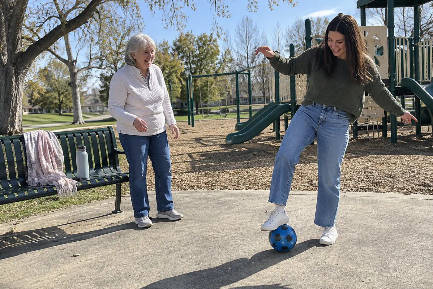 A grandmother watches her granddaughter roll a ball beneath one shoe beside a playground bench.