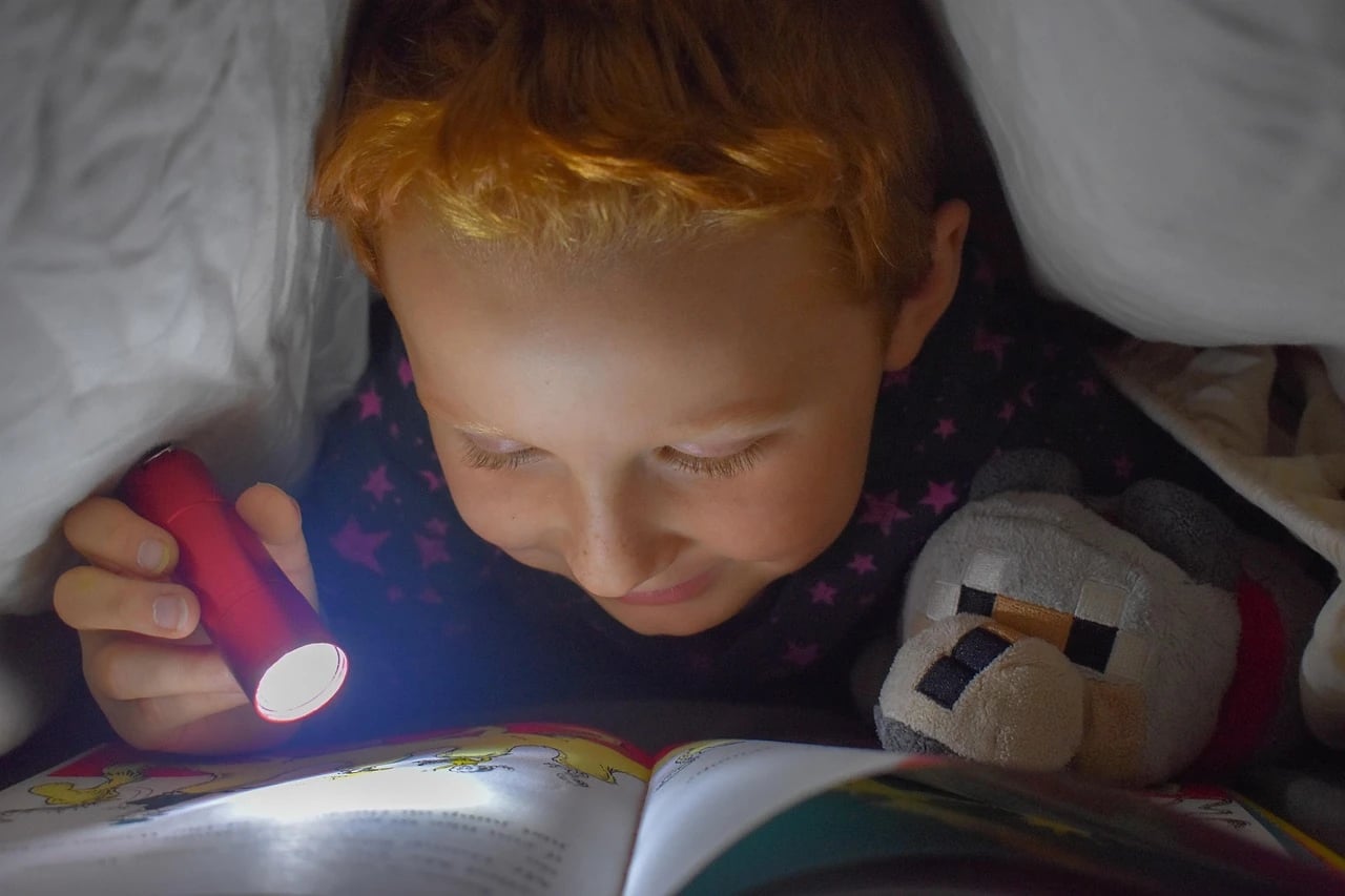 Red-haired child reading a picture book by flashlight under a blanket with a plush toy beside him