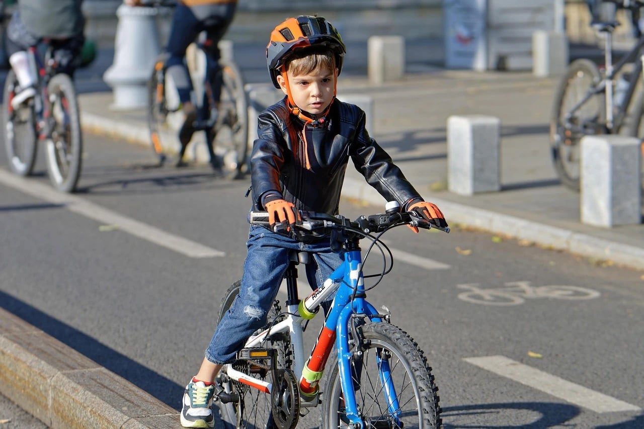 Young boy in an orange helmet and leather jacket rides a blue bicycle along a city bike lane