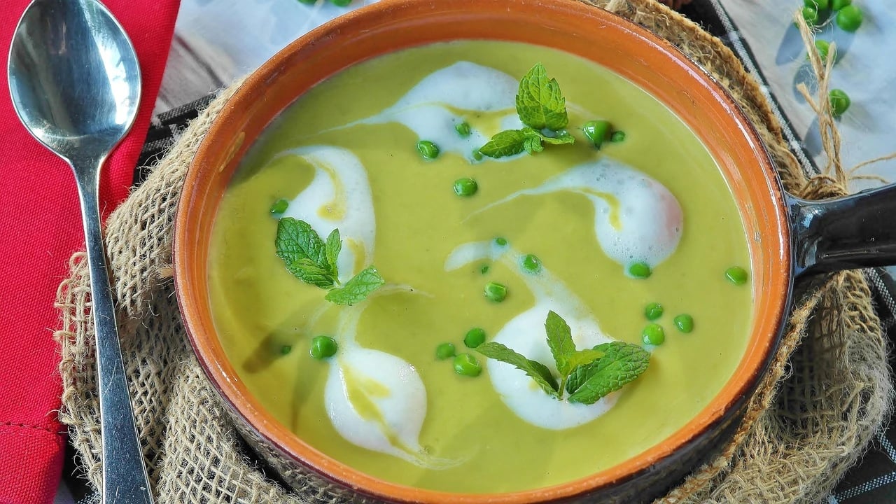Bowl of green pea soup with cream swirls, mint leaves and whole peas on burlap beside a spoon