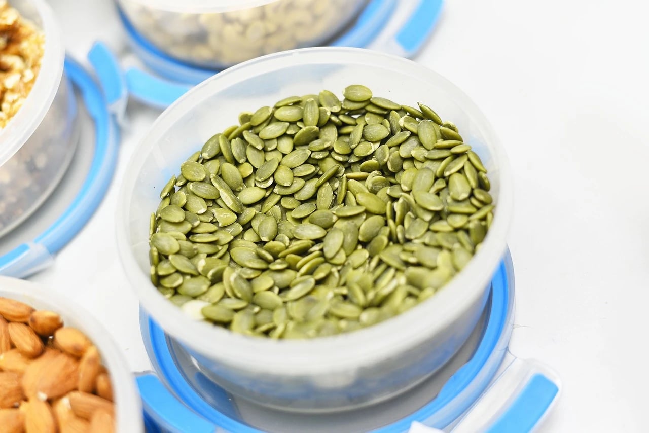 Green hulled pumpkin seeds in a clear plastic container with blue lid, surrounded by almonds and other nuts