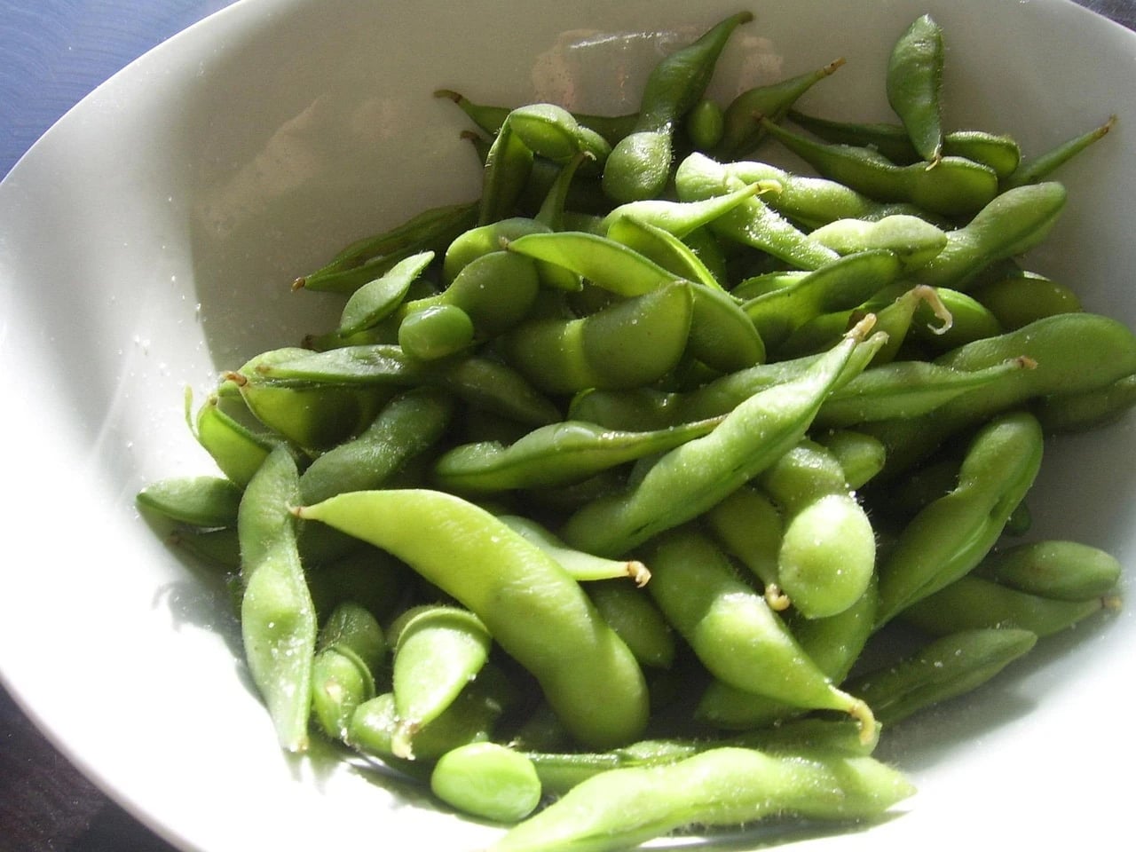 White bowl filled with green edamame pods lit by sunlight, lightly salted, on a dark table