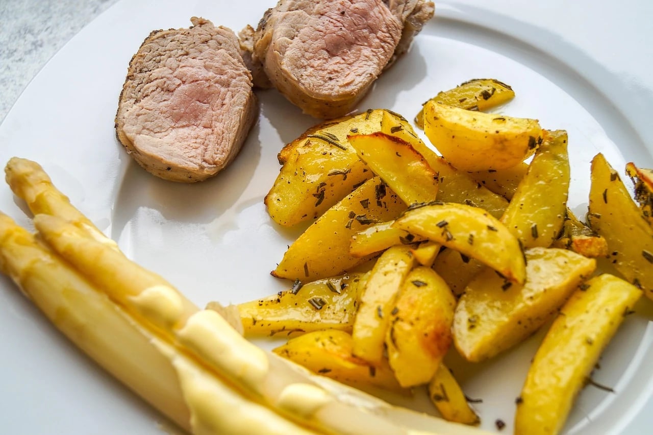 Sliced pork tenderloin medallions on a white plate with rosemary roasted potatoes and white asparagus with hollandaise