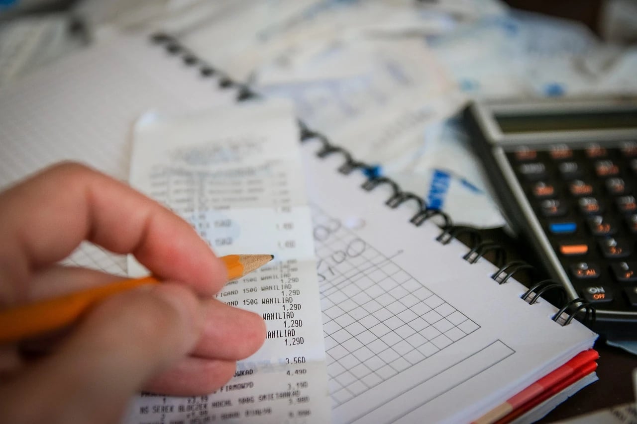 Hand holding a pencil over a store receipt beside a spiral notebook and calculator while working on a budget