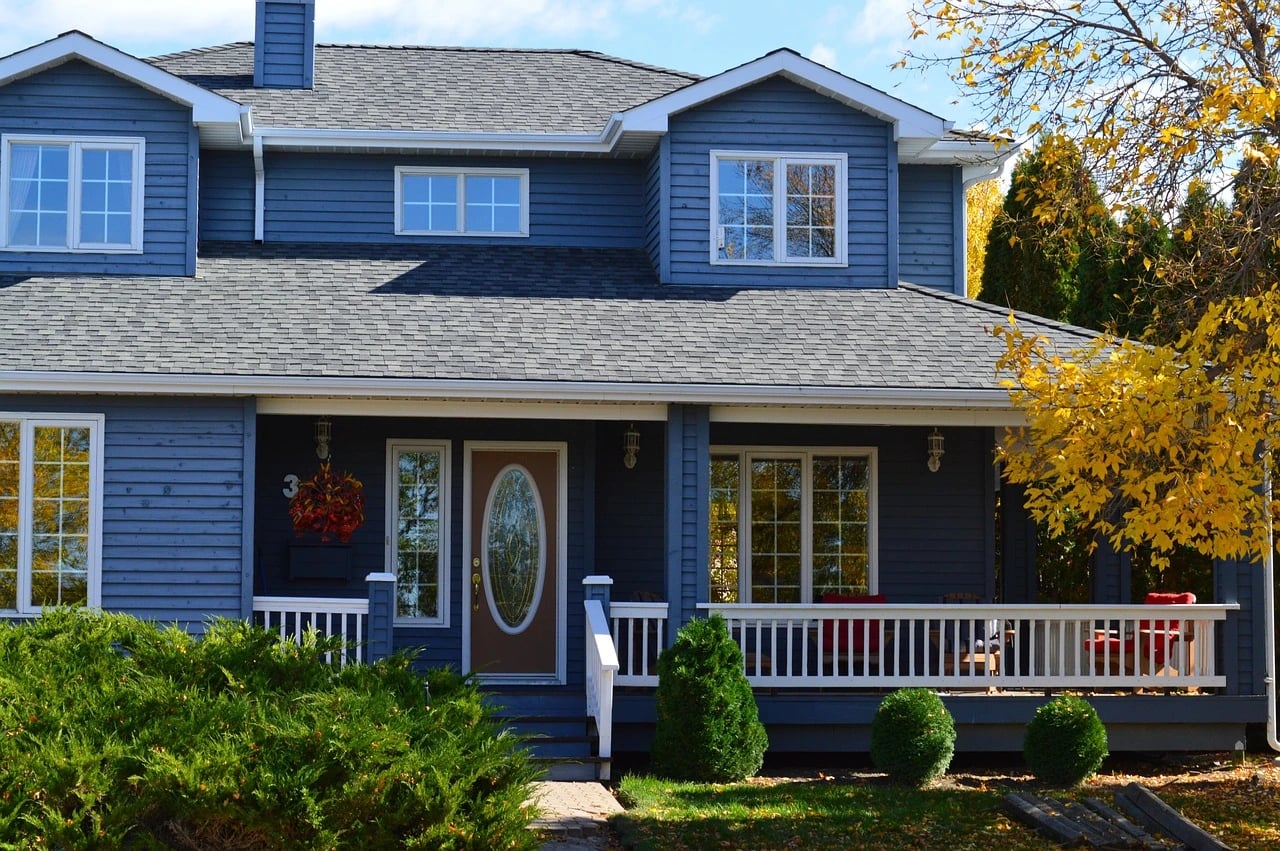 Blue two-story house with white trim, covered front porch, shrubs and yellow autumn tree on a sunny day