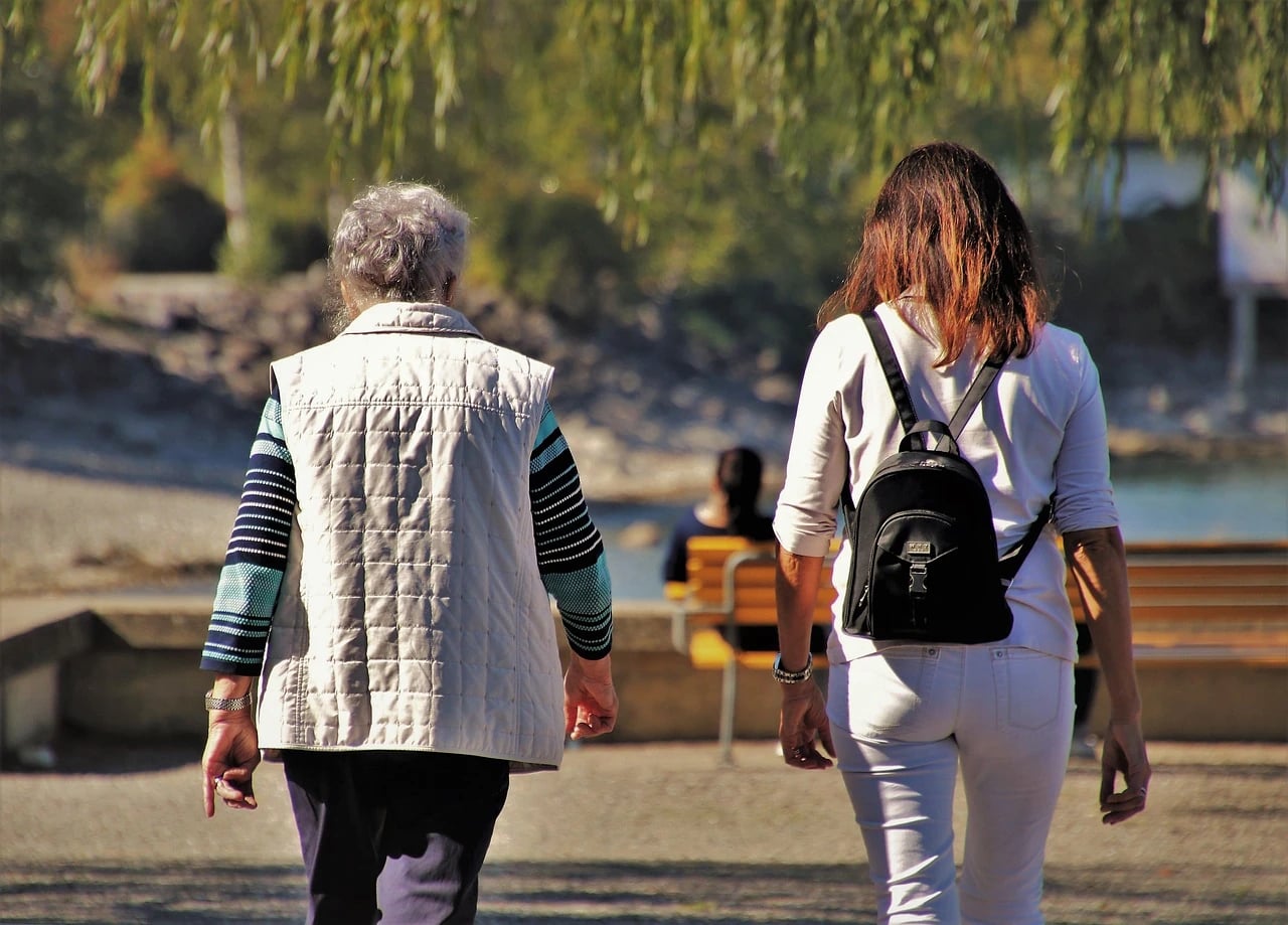 Two women seen from behind walking together along a sunny lakeside park path past a wooden bench