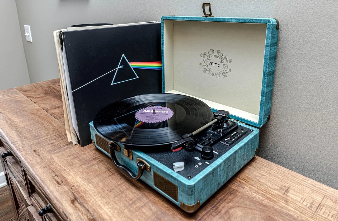 Teal portable record player with a vinyl record playing beside a stack of albums on a wooden dresser