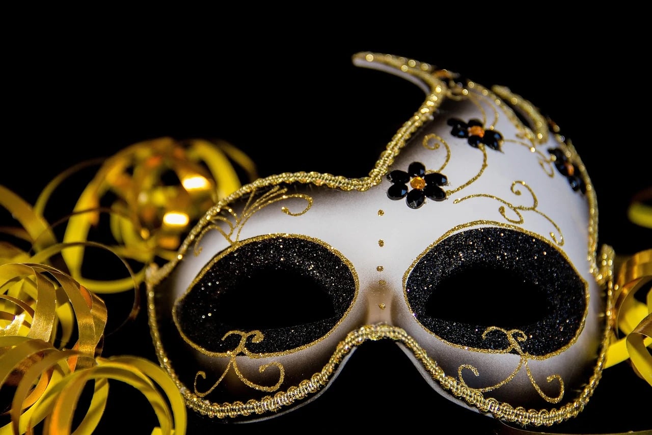 White and gold glittery masquerade mask with black flowers beside curling gold ribbons on a black background