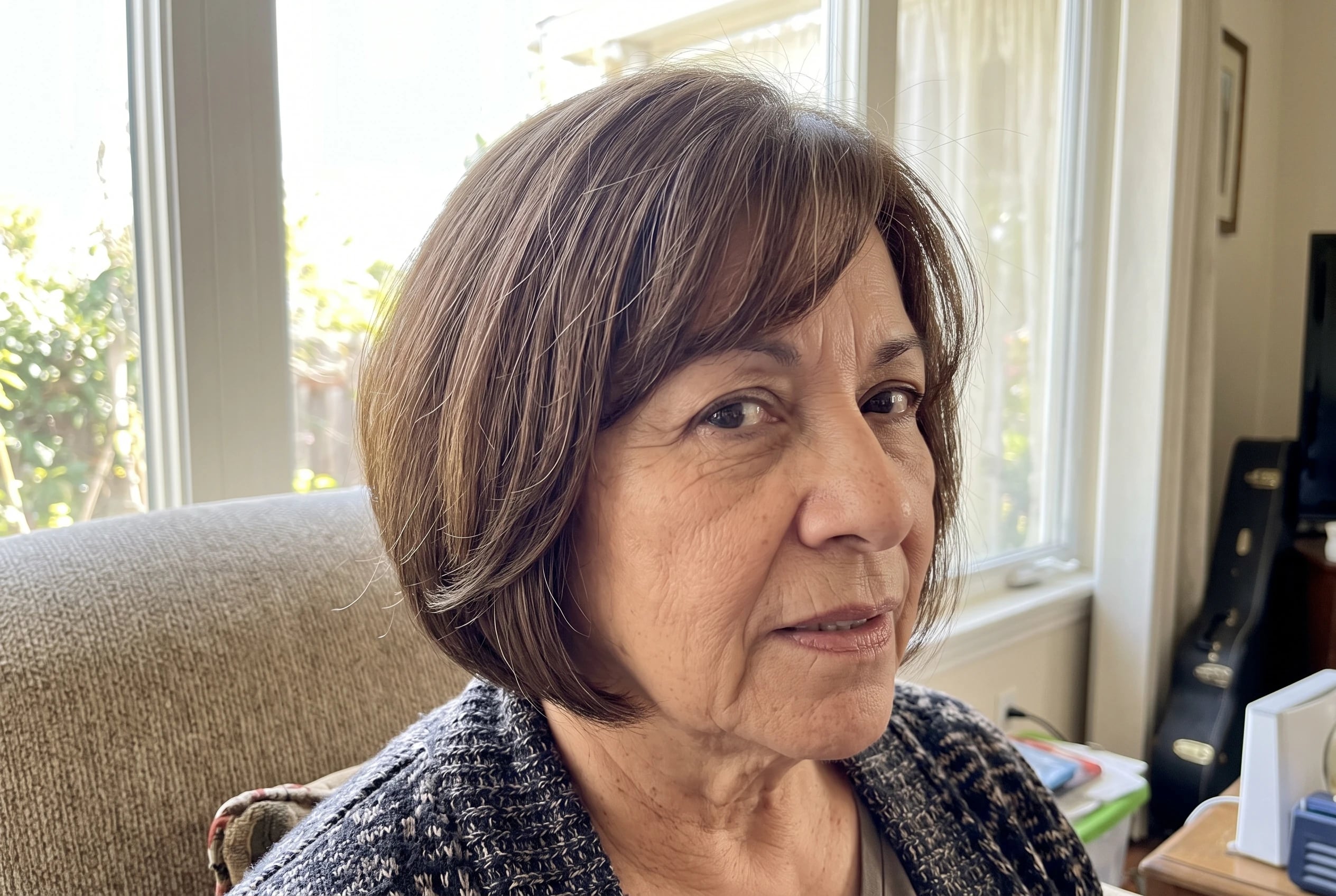 A rounded bob with side bangs curves around the cheek of a woman in her sixties seated by a bright living-room window.
