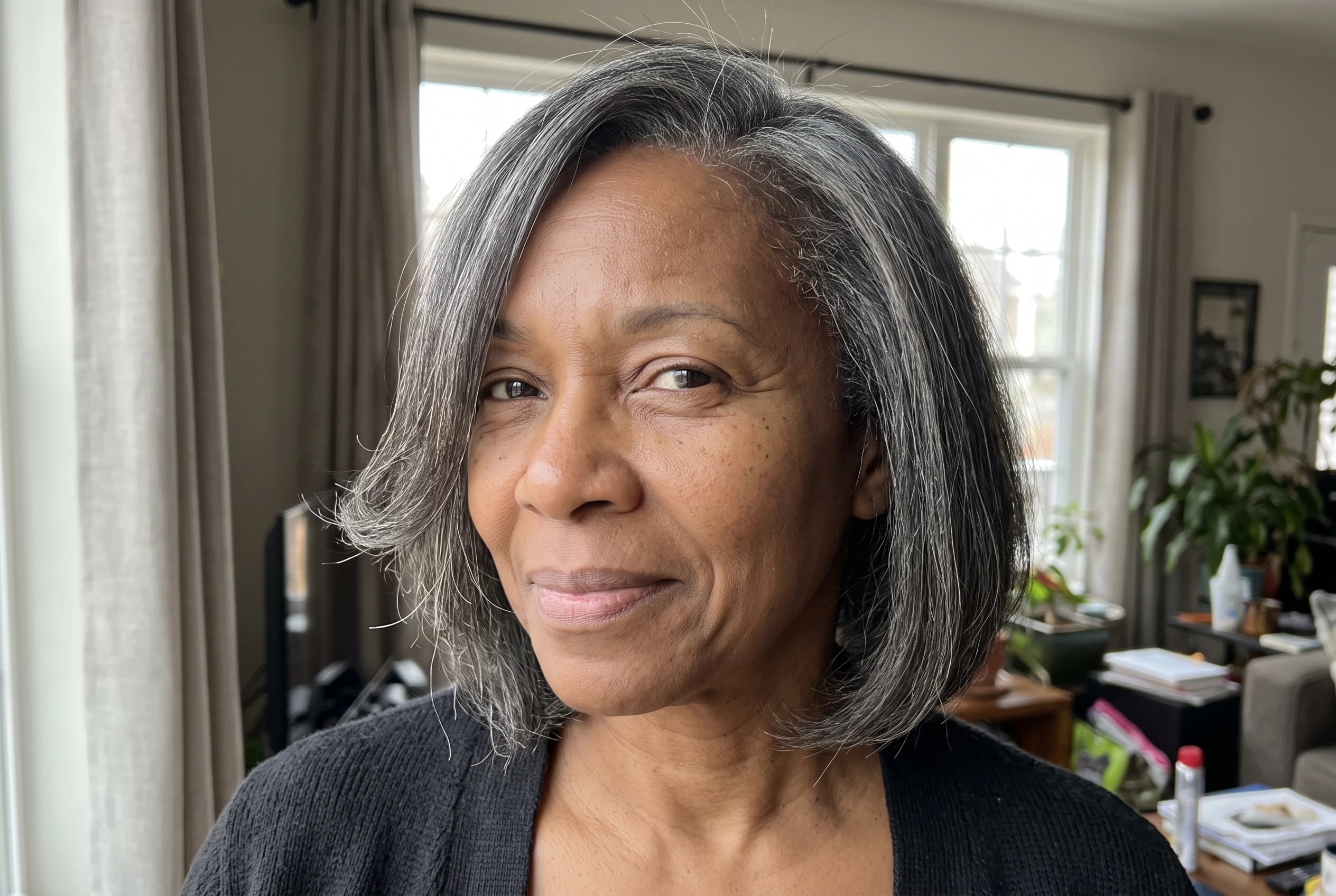 A chin-length bob with a deep part lifts on one side as a woman in her fifties stands beside a living-room window.
