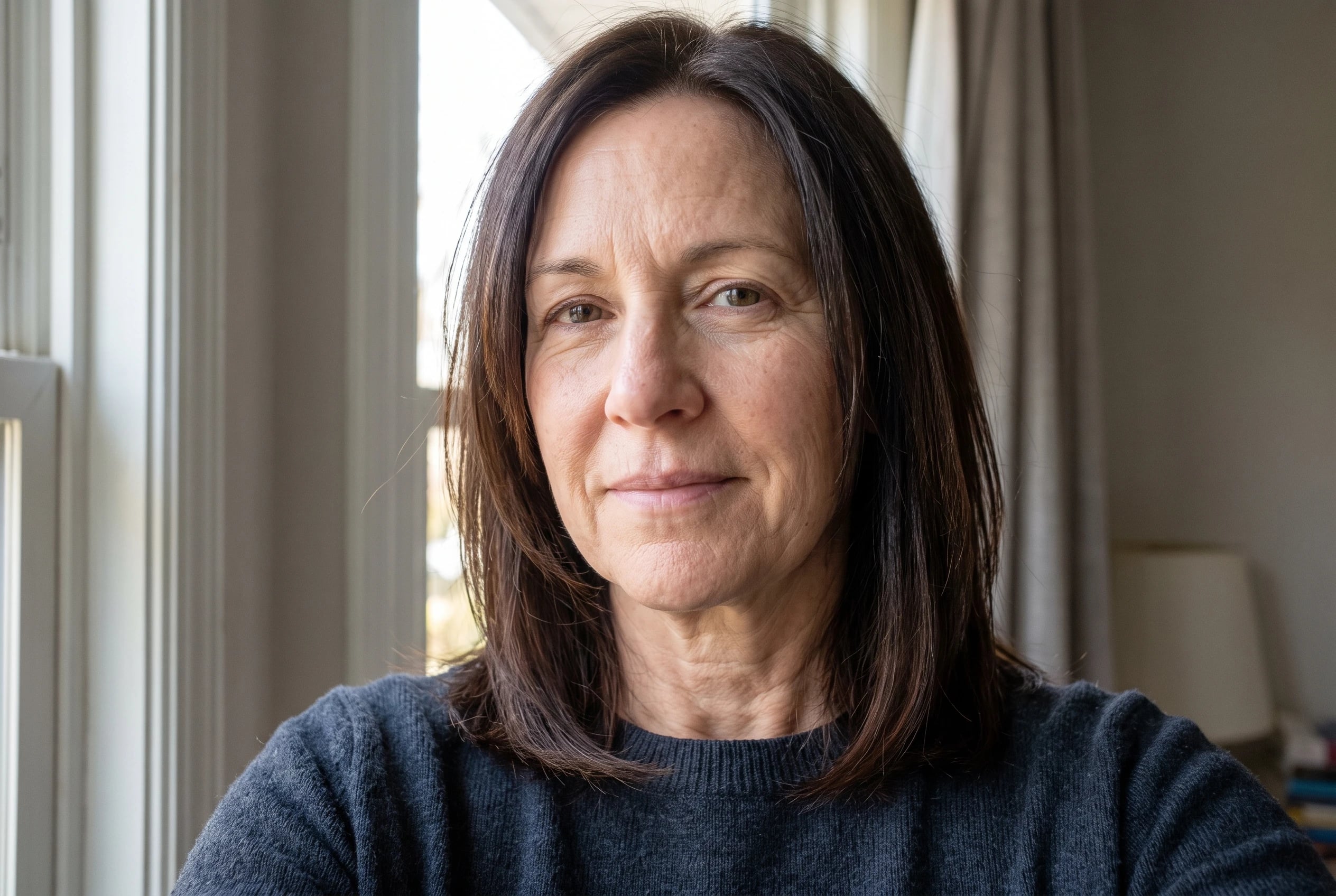 A collarbone cut with long layers lies over a dark sweater as a woman in her fifties stands near a bedroom window.