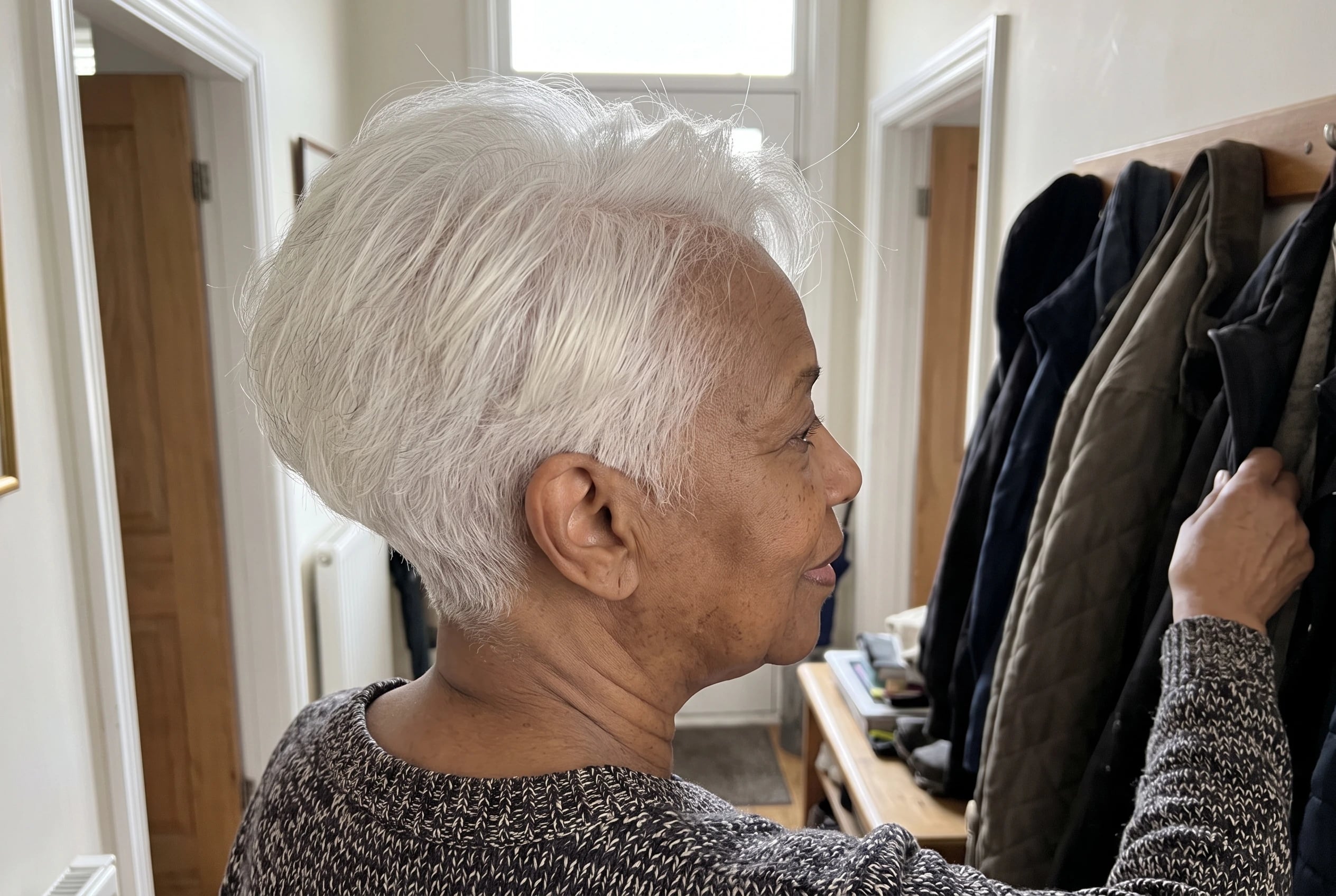 A tapered nape with a full crown is seen from behind as a woman in her seventies reaches for a coat in her hall.