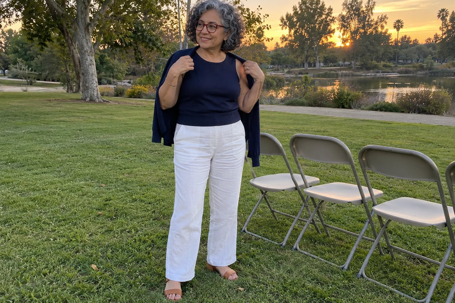 A woman in her sixties stands by folding chairs on a lawn at sunset, wearing white linen trousers, a navy knit top, cardigan draped over her shoulders, red glasses and tan sandals.