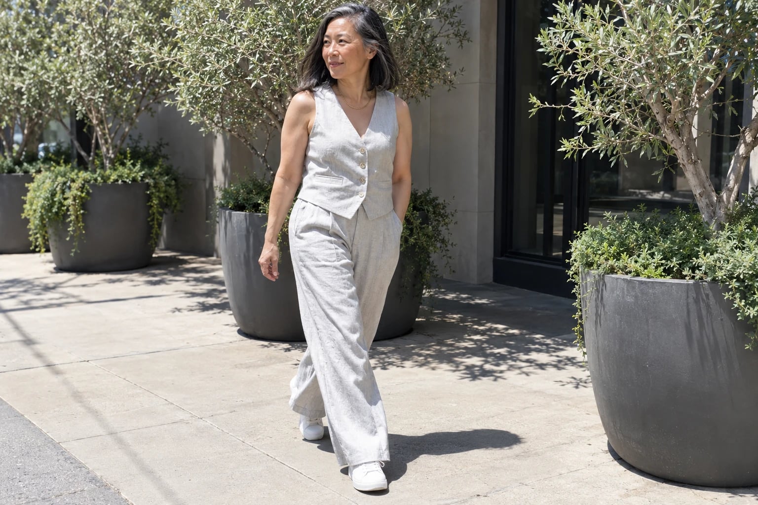 A woman in her fifties walks past potted olive trees in a buttoned pale gray linen vest and wide-leg trousers.