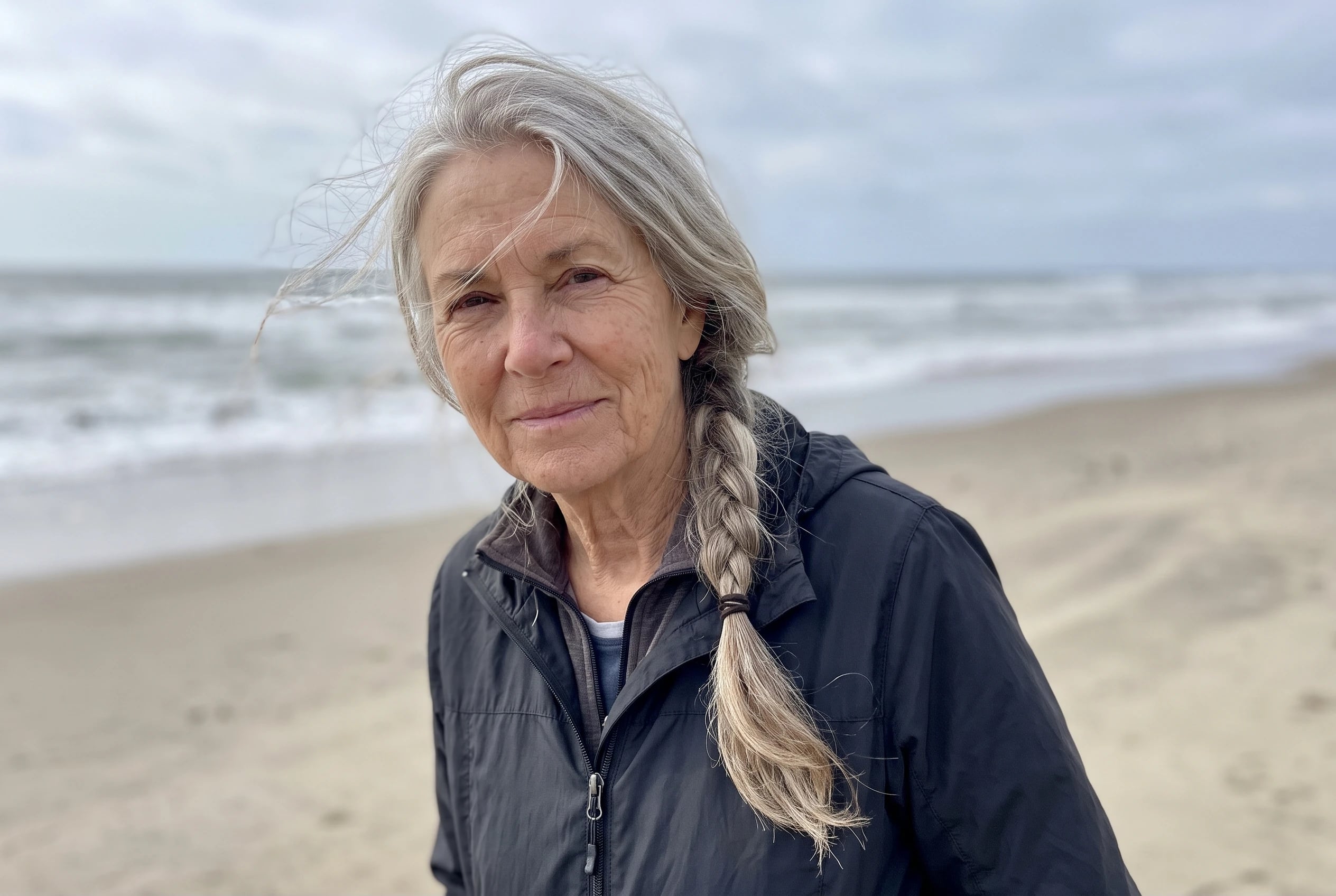 A side braid with loose roots falls across the shoulder of a woman over 50 walking on a windy beach.