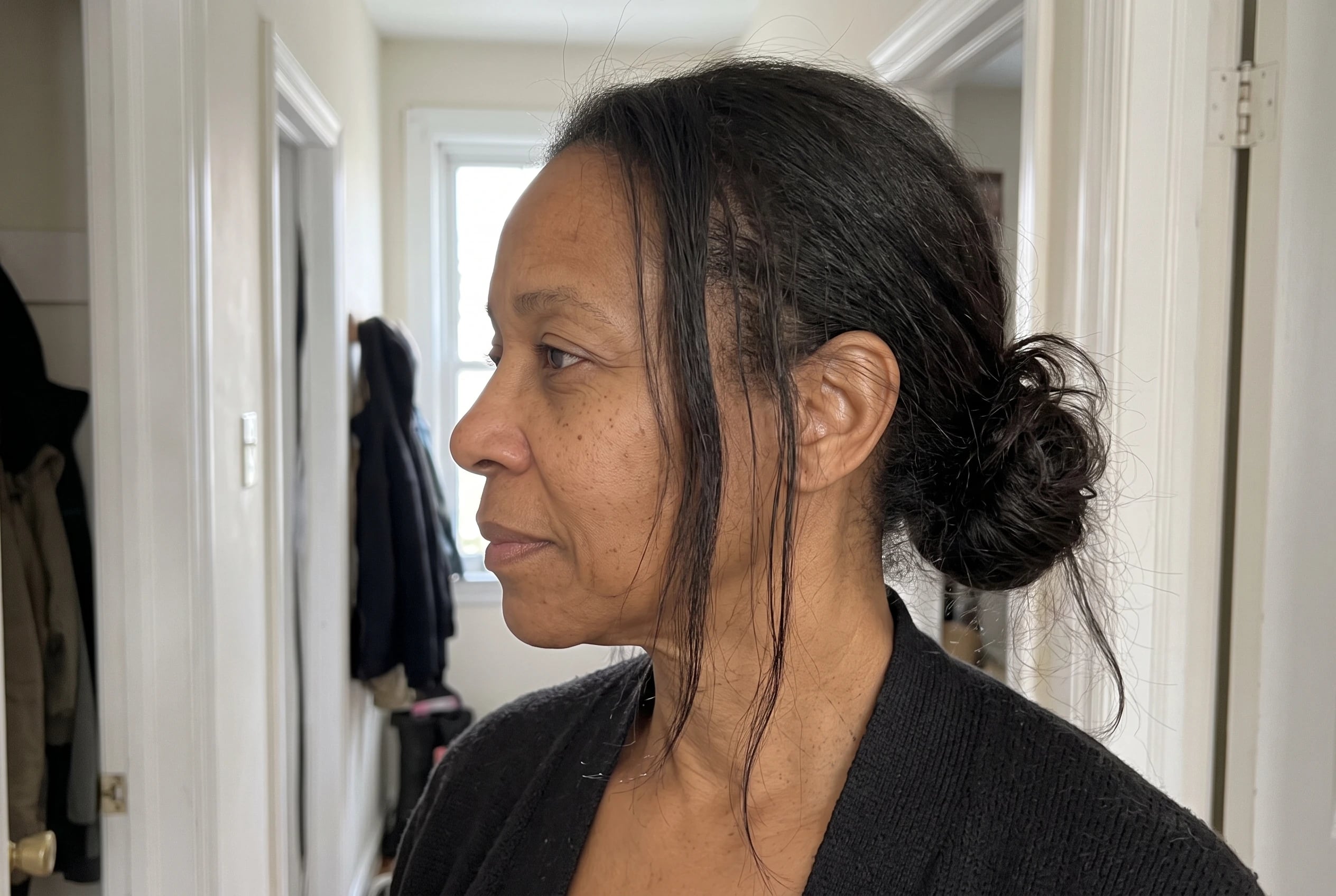 A low bun with loose lengths frames a woman in her late fifties as she fastens her coat in a hallway.