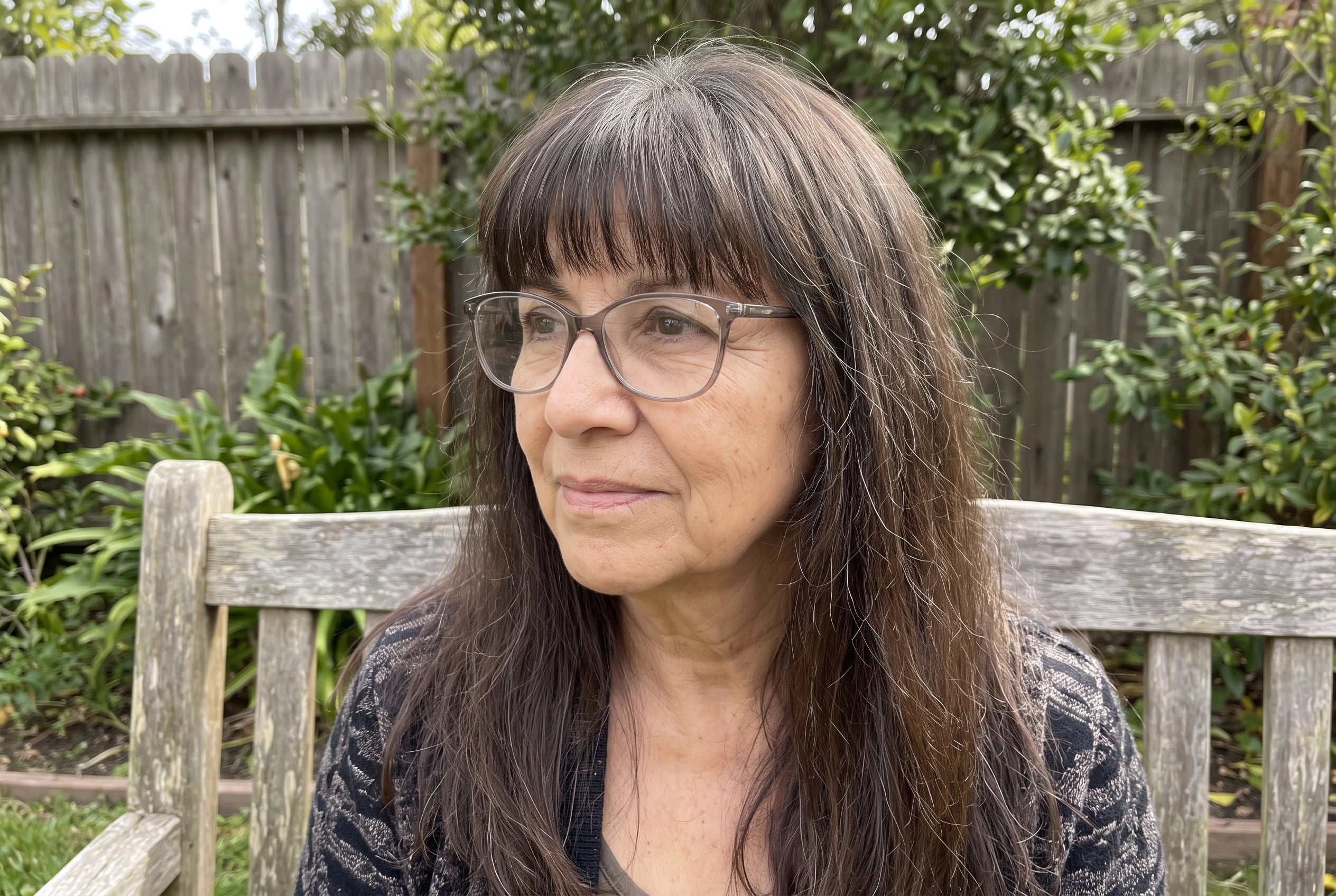 Long hair with a full fringe frames a woman in her sixties wearing glasses as she sits on a garden bench.
