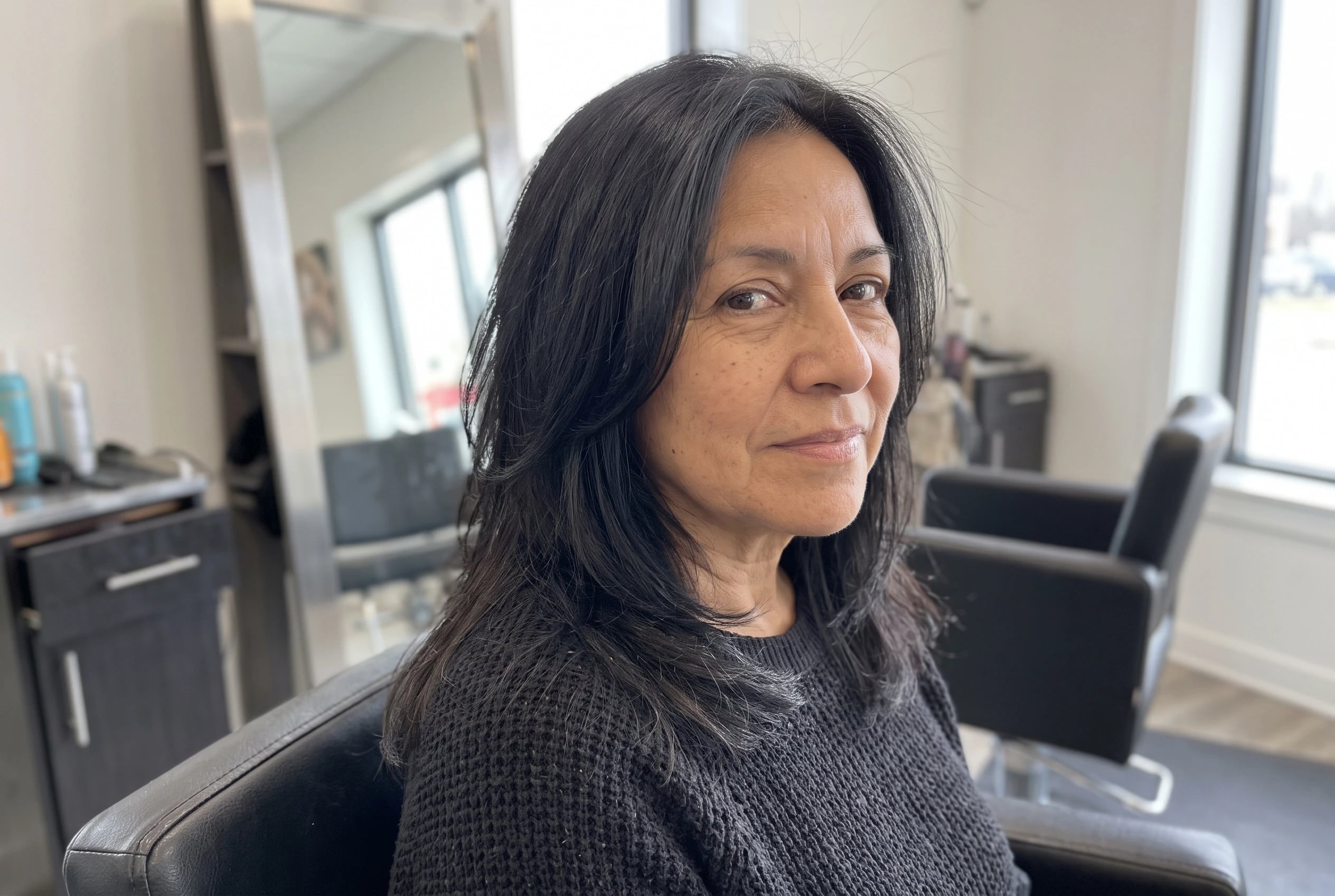 Long layers around the collarbone frame a woman in her late fifties as she turns her head in a salon chair.