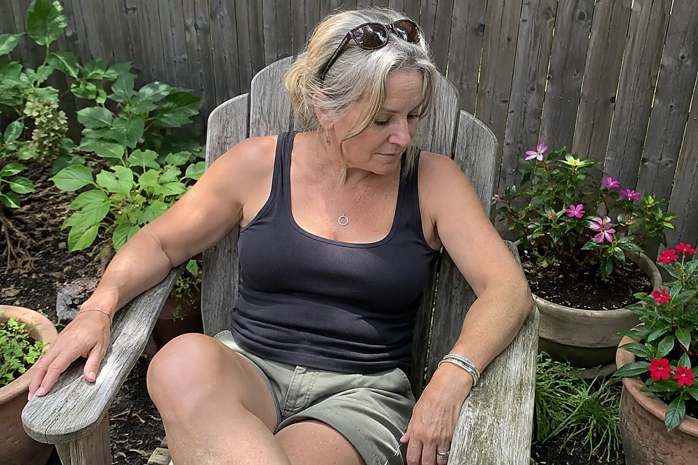 A woman in her fifties sits in a wooden chair among potted garden plants.