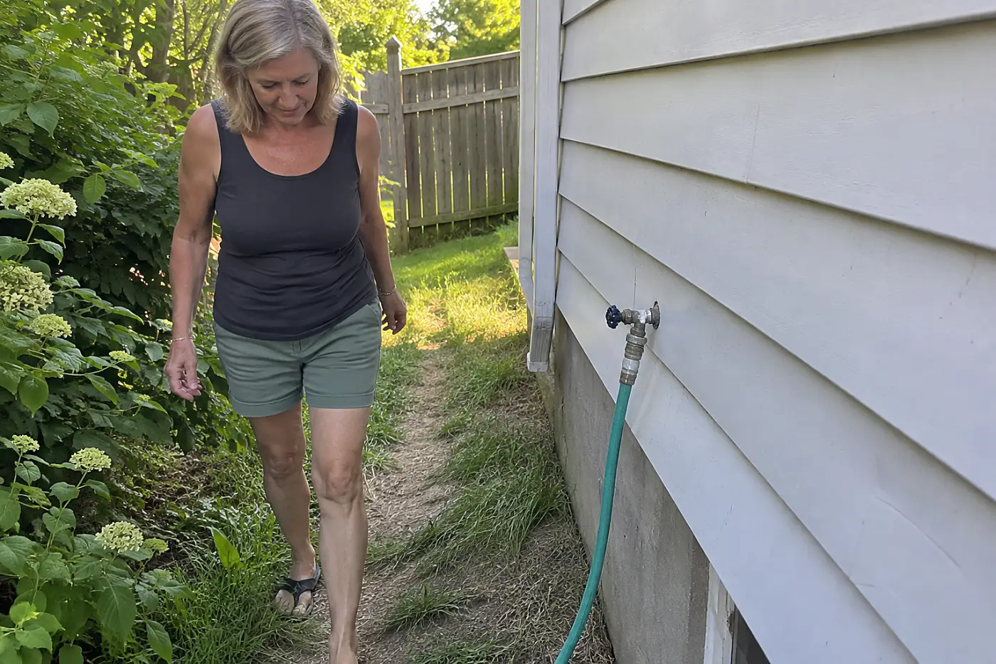 A woman in her sixties walks along a worn path through grass beside a house.