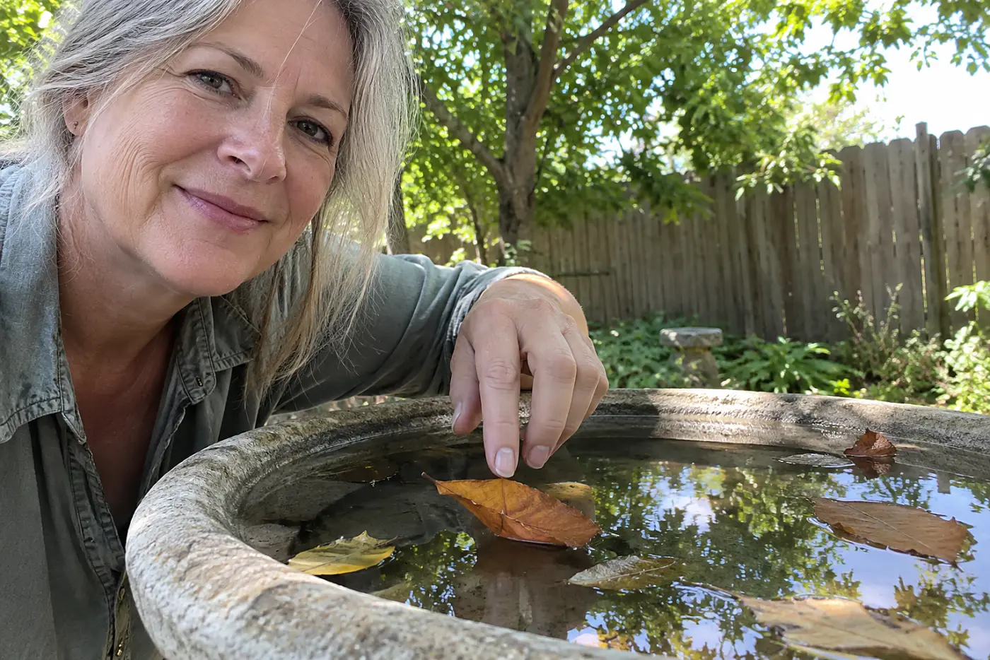 A smiling woman with grey hair touches a leaf floating in a garden birdbath.