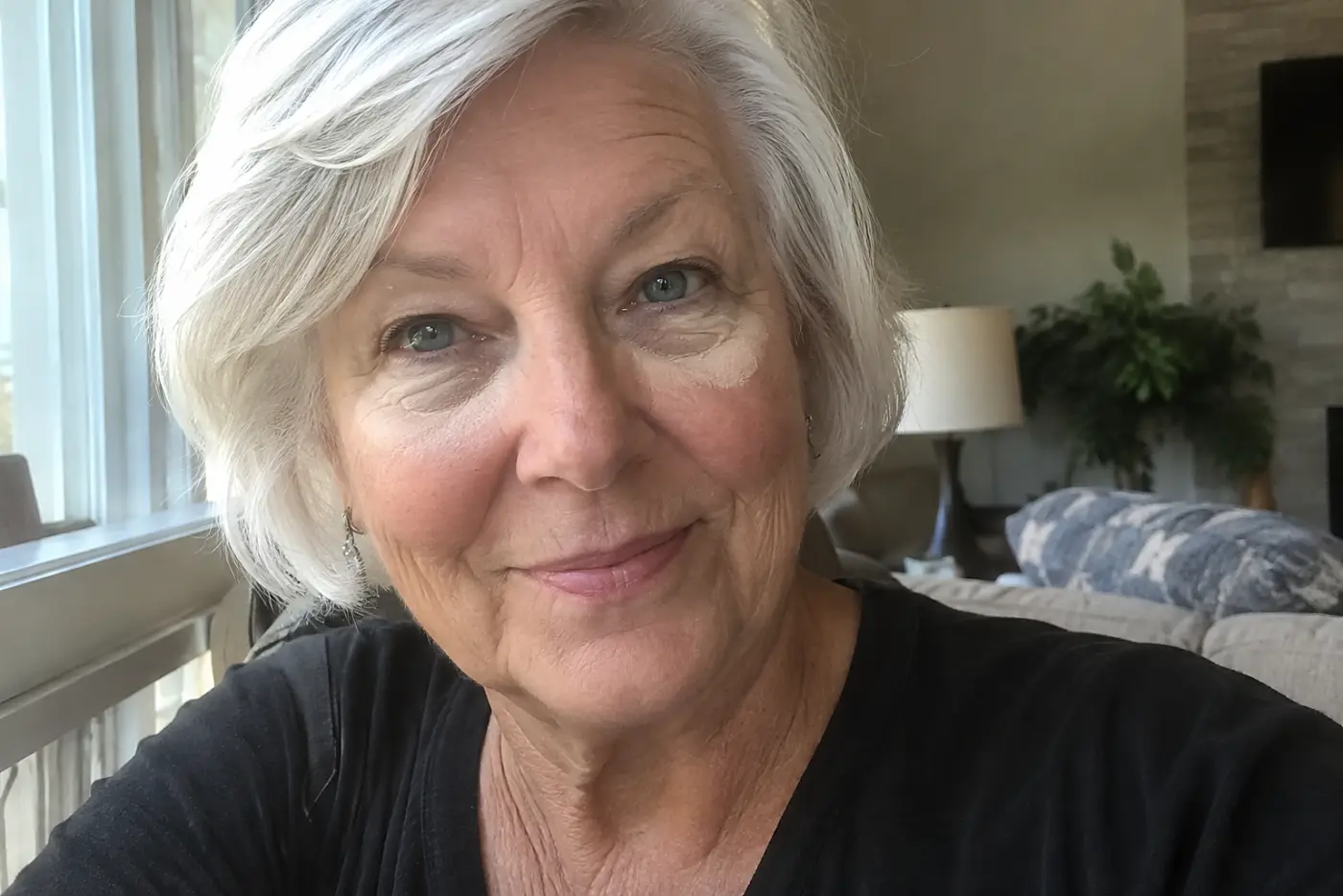 A woman in her sixties with pale powder under her eyes sits near a window.