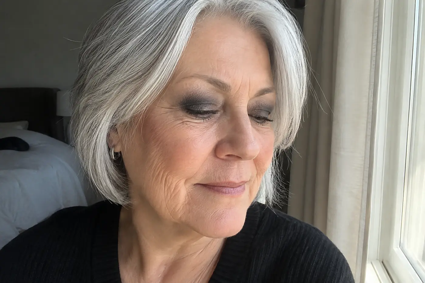 A woman in her sixties with dark eyeshadow in her crease looks toward a window.