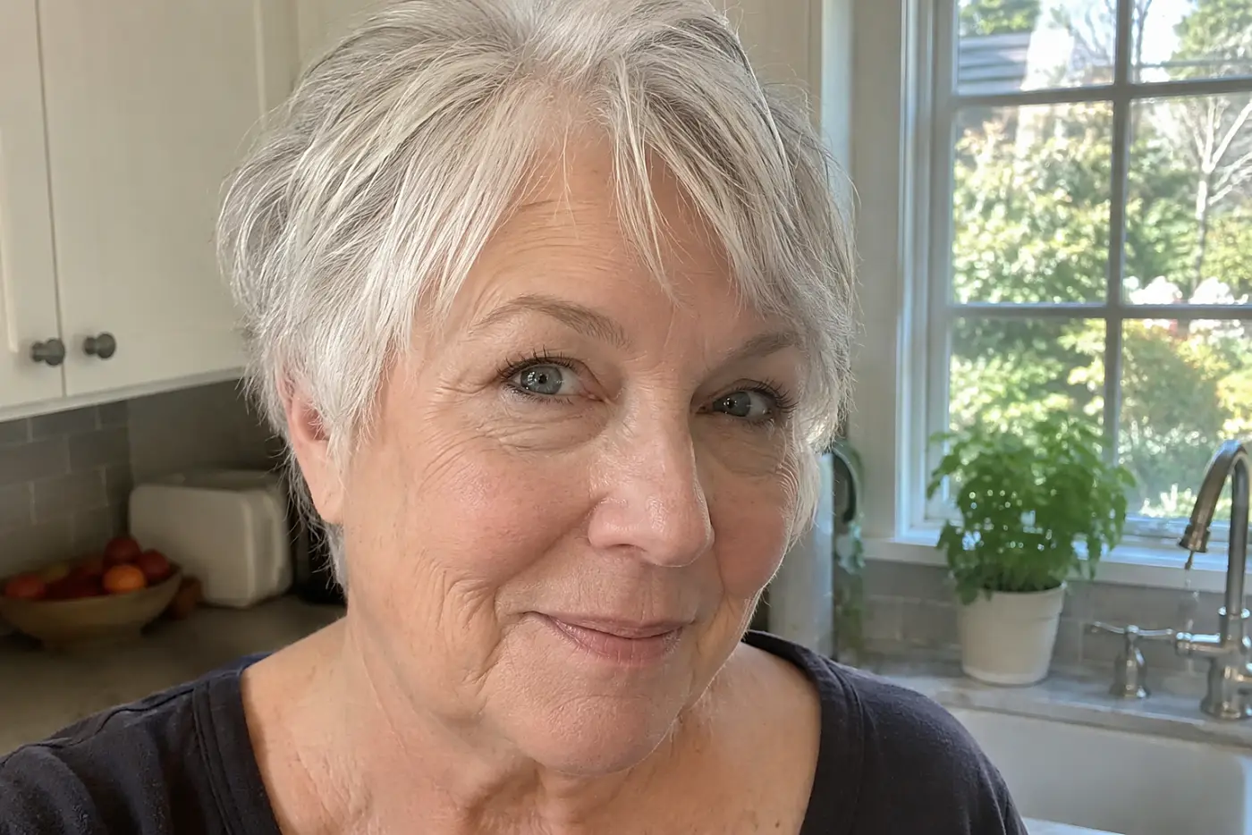 A woman in her sixties with short grey hair smiles beside a sunlit kitchen window.