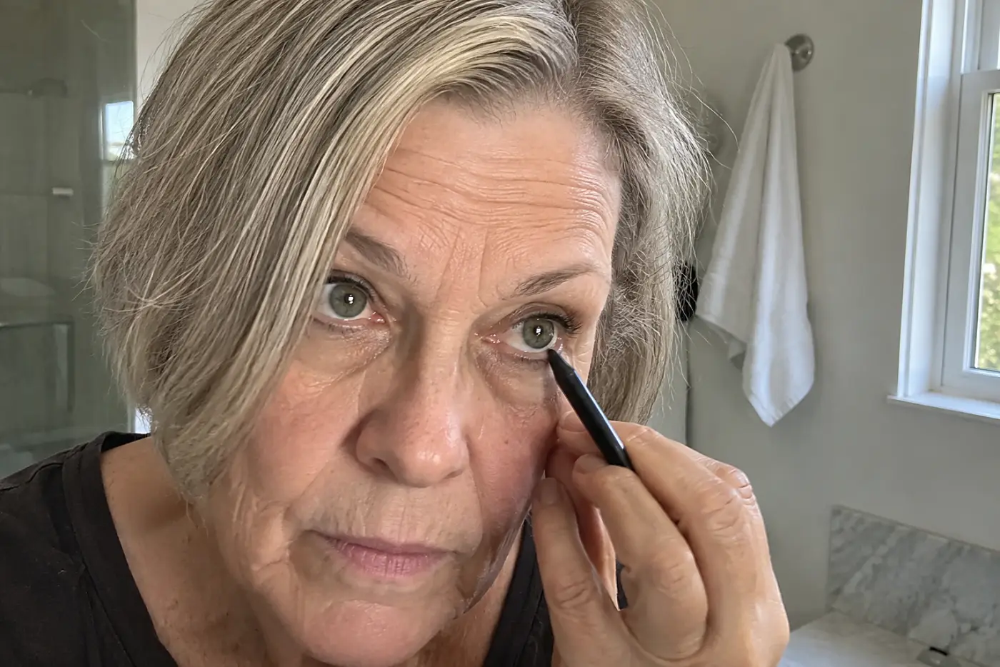 A woman in her sixties applies black eyeliner to her lower lash line.