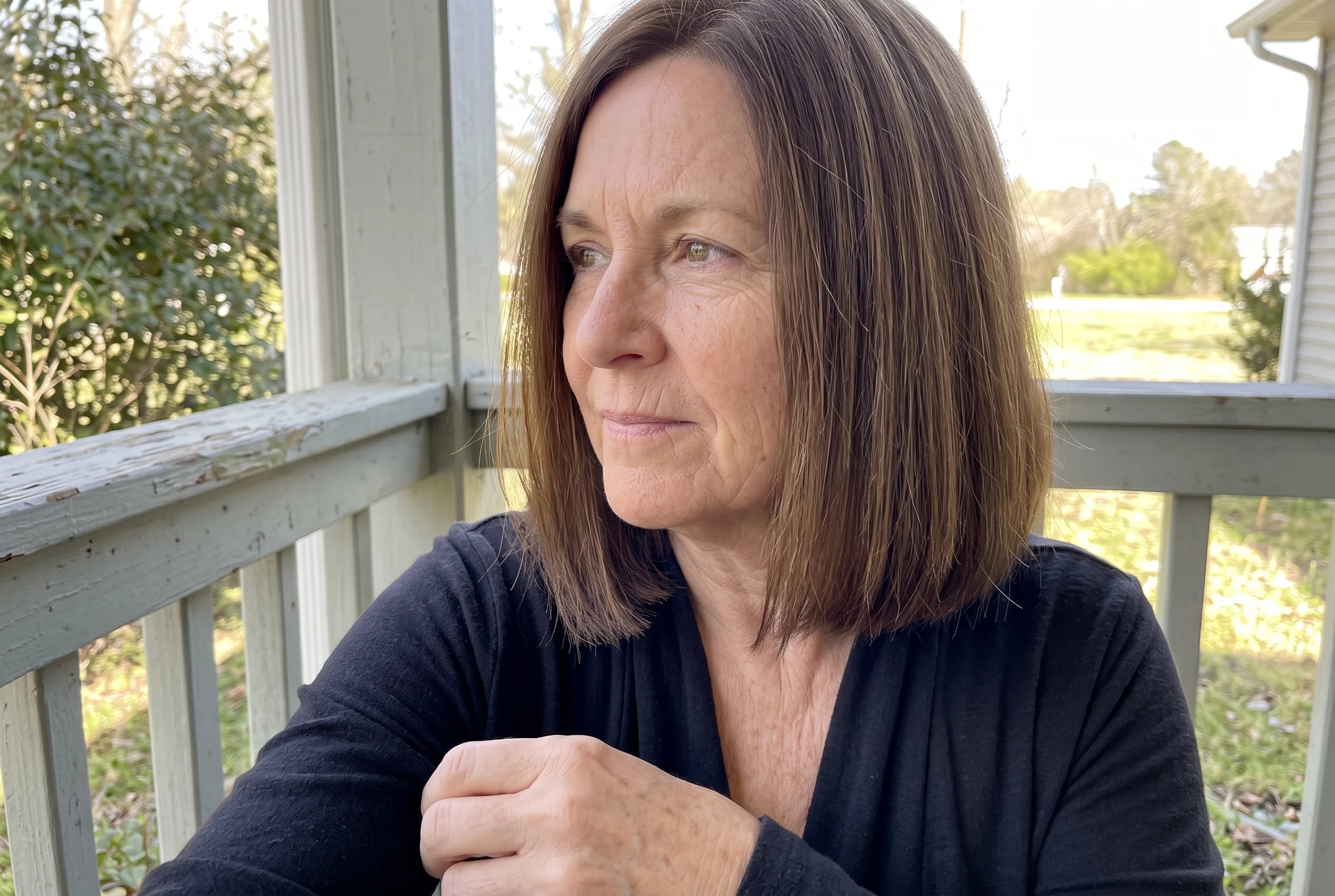 A woman in her sixties with a brown blunt lob hairstyle looks out from a porch.