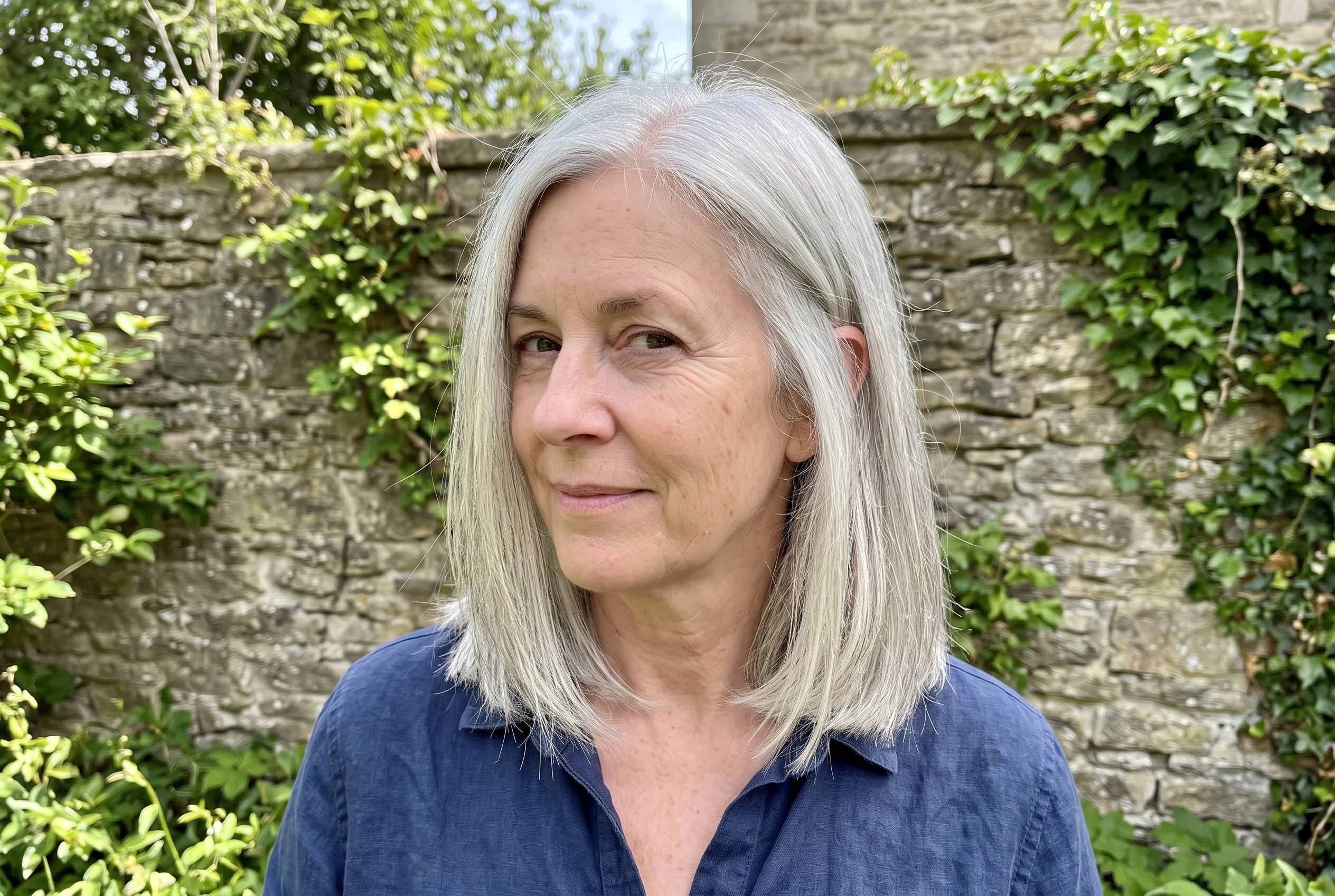 A woman in her sixties displays a silver lob tucked behind one ear near a garden wall.