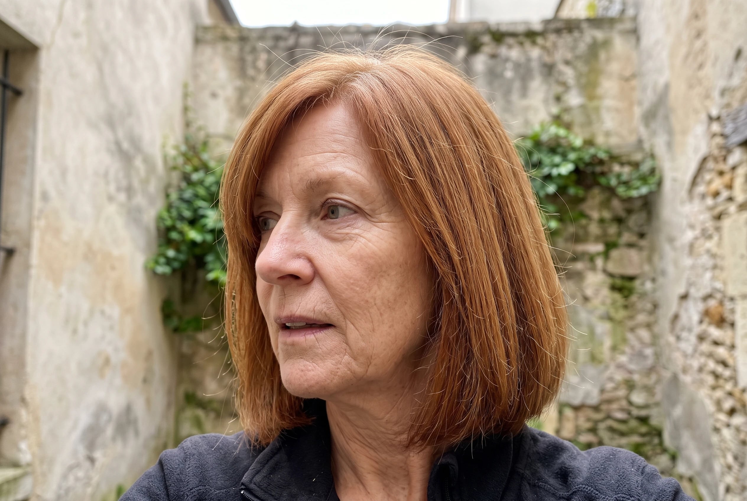 A woman in her sixties with mid-length red hair stands in a stone courtyard.