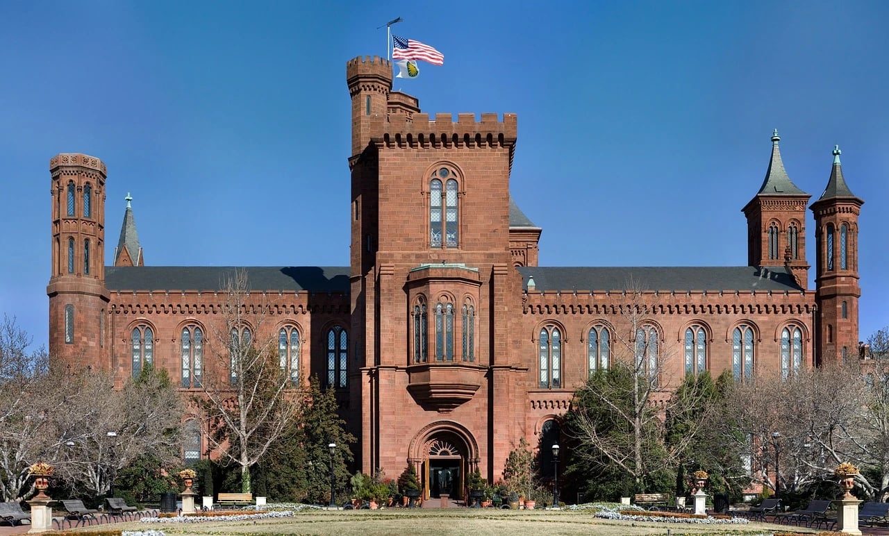 Red sandstone Smithsonian Castle with towers and an American flag, seen across a garden under blue sky