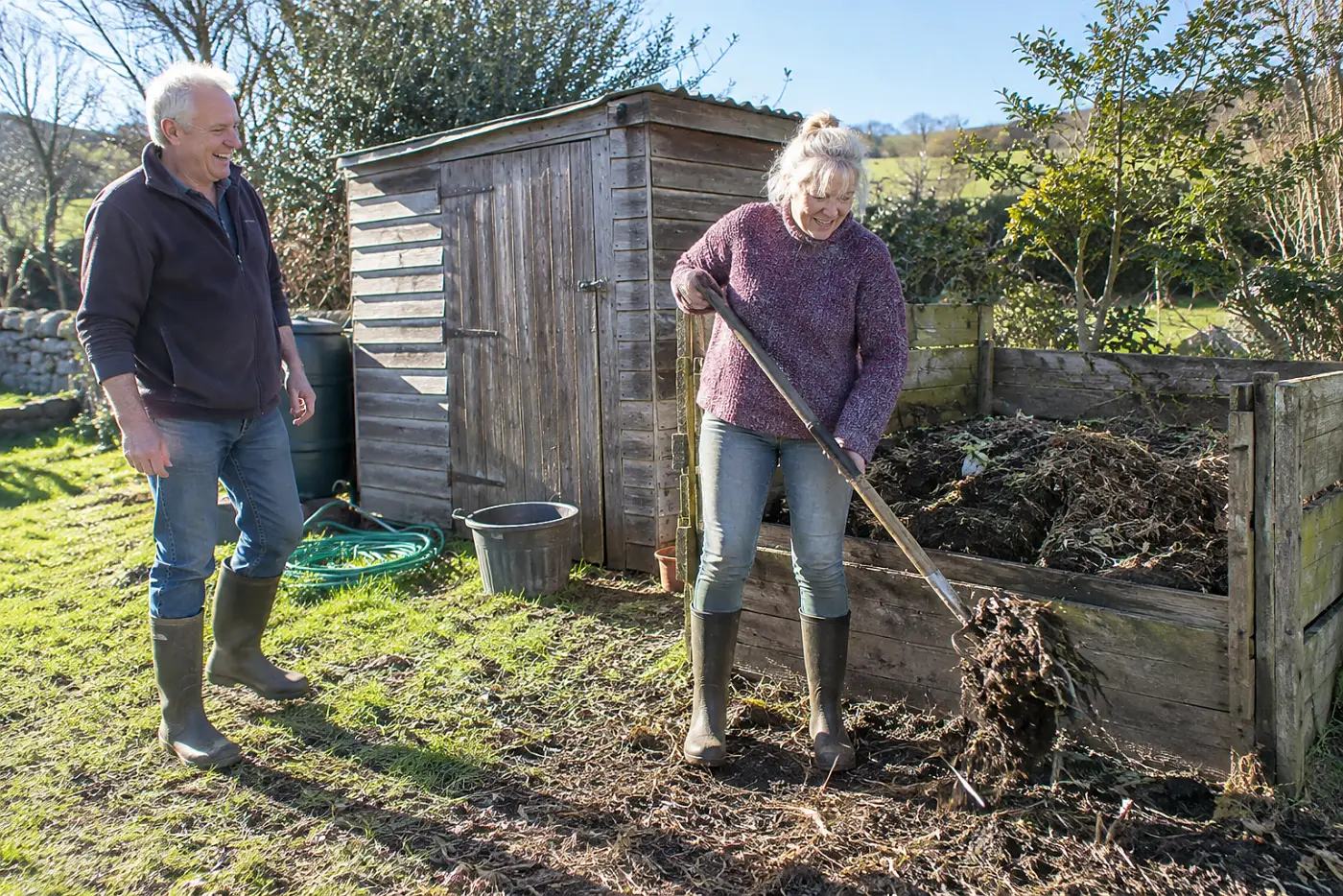 Older woman turning a compost pile with a fork behind a shed.