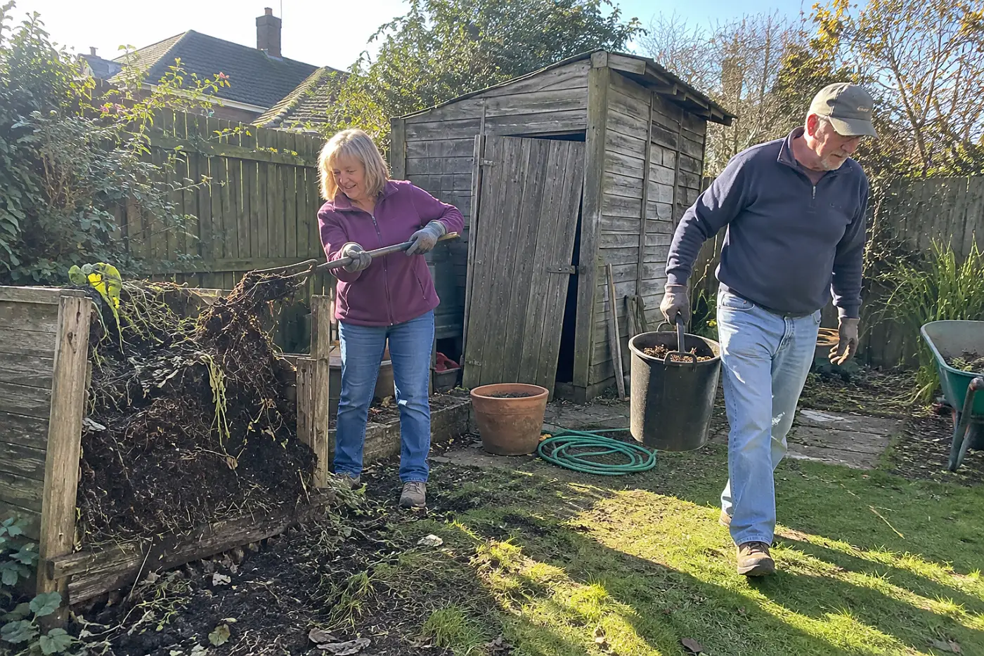 Older woman turning a compost pile with a garden fork behind her shed.