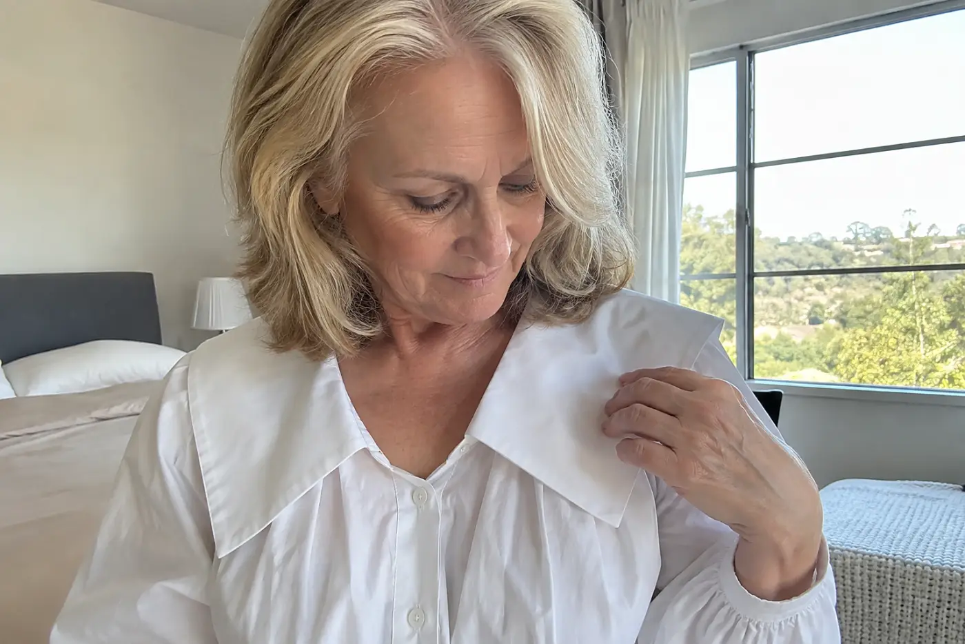 A woman in her sixties touches the large collar of her white blouse.