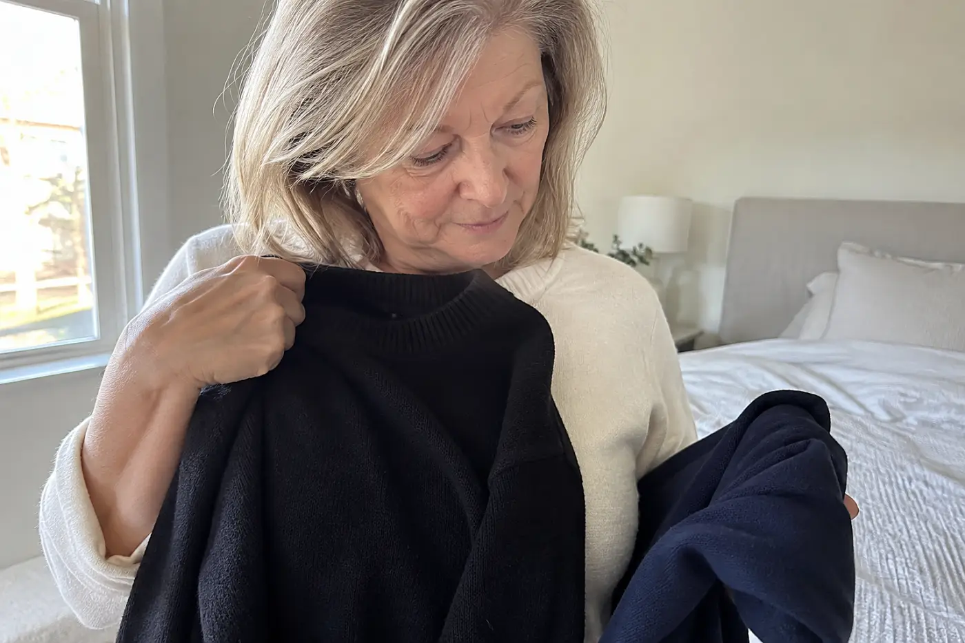 A woman in her sixties holds up a black sweater beside a navy shirt.