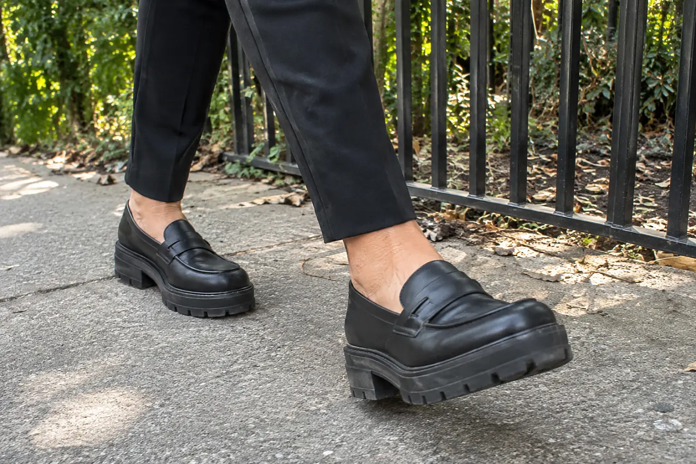 A woman in her sixties walks along a sidewalk wearing dark trousers and heavy-soled shoes.