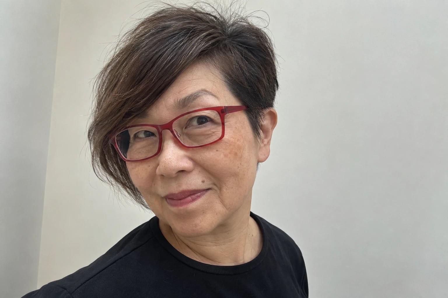 A woman in her sixties wears a dark asymmetrical pixie cut and red rectangular glasses, smiling softly against a plain wall.