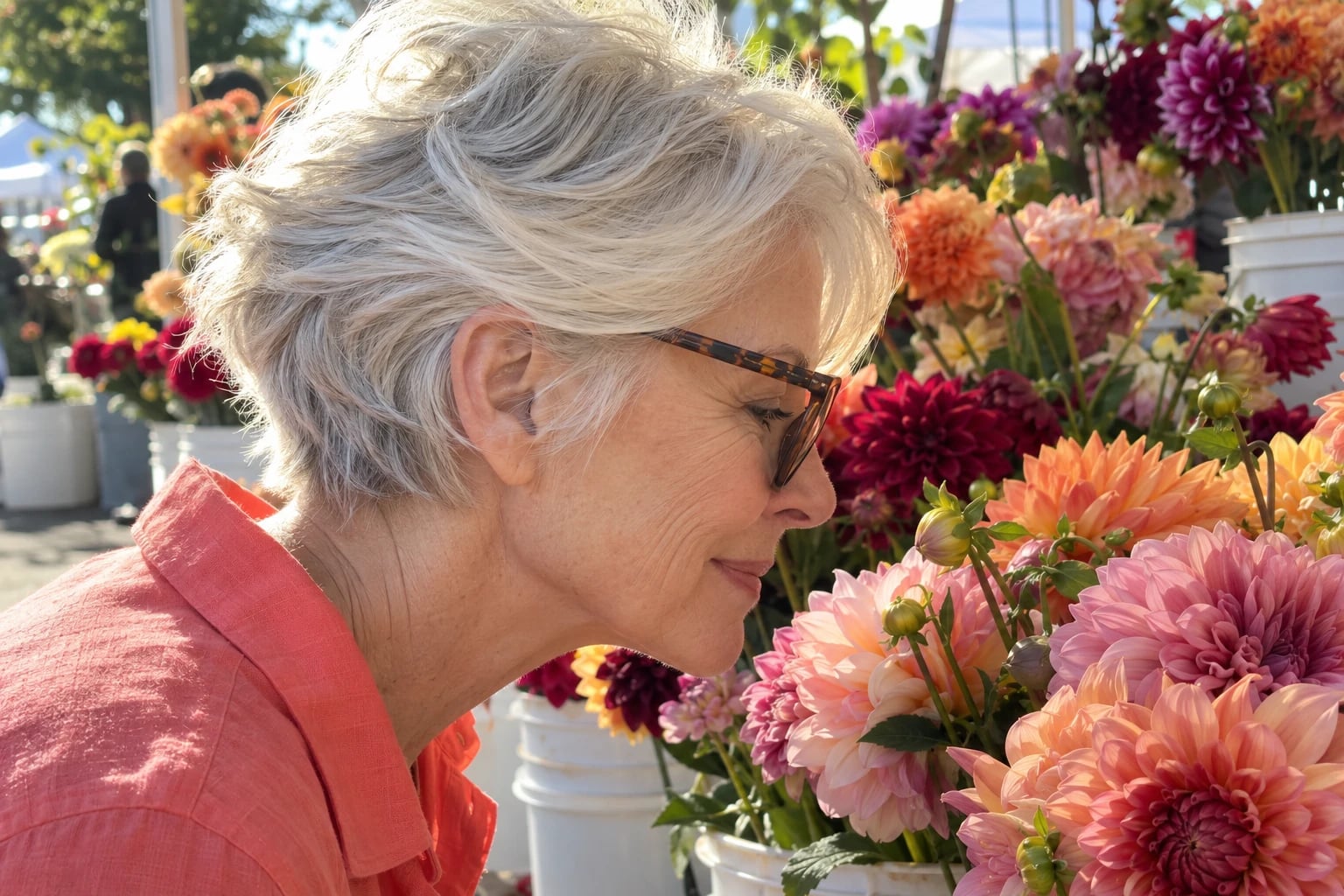 A woman in her sixties with a silver feathered pixie and tortoiseshell glasses smells colorful dahlias at a market stall.